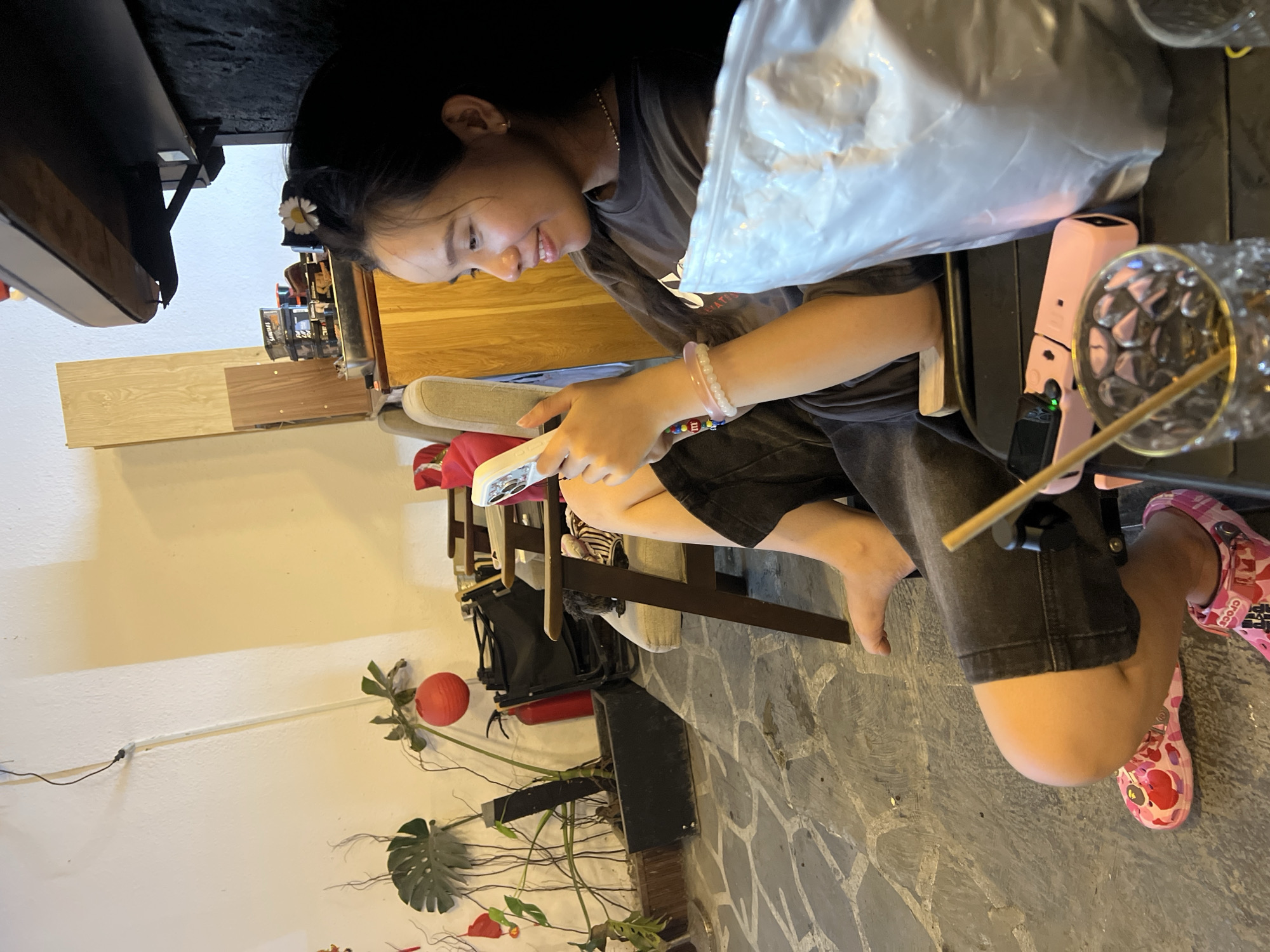 An indoor shot of a young woman sitting at a table in what appears to be a café. The location is presumed to be in Đà Nẵng, Vietnam. The woman has dark hair, a flower clip, and is wearing a gray t-shirt and dark shorts. She is holding a smartphone and looking at it with a smile. On the table are a disposable plastic bag, a pink electric outlet box, and a glass with a straw. She wears a bracelet on her left arm. On her feet are pink Crocs. The surrounding environment includes a stone floor, a wooden chair, and a potted plant in the corner. Behind the woman, there is a wooden wall and what appears to be shelves with items on them.