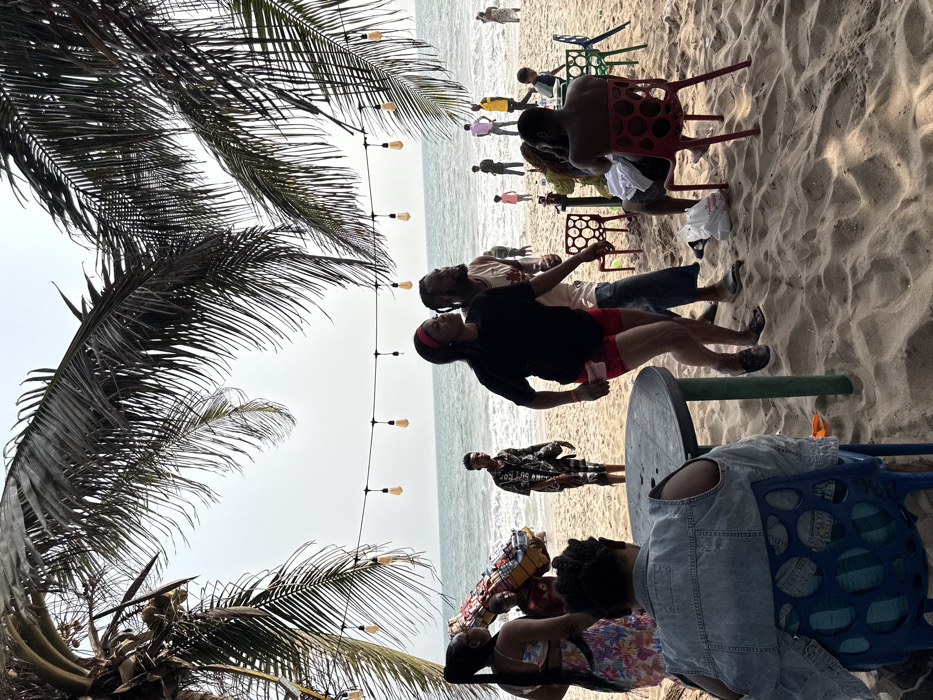 The scene depicts a beach environment in Mopo Onijebu, Nigeria, under an overcast sky. The foreground features a sandy beach with a person seated on a blue plastic chair, their back to the camera, wearing a denim vest. A dark, round plastic table is positioned directly in front of this individual.
To the left, a woman in a floral two-piece garment stands, observing a person with merchandise balanced on their head. Further towards the water, a man in a black and white patterned ensemble stands at the edge of the ocean.
In the mid-ground right, a woman in a black short-sleeved top, red shorts, and a red headband moves or dances towards the right, alongside a man with dreadlocks wearing a patterned shirt and jeans. Nearby, a shirtless man sits on a red plastic chair, facing away from the camera. Another person is seated on a green plastic chair.
A string of incandescent light bulbs is suspended above the mid-ground. The background reveals the ocean with small waves and numerous individuals scattered along the shoreline and within the shallow water. Tall palm fronds frame the upper portion of the image, indicating their presence overhead. The overall setting suggests a casual gathering or leisure activity by the sea.