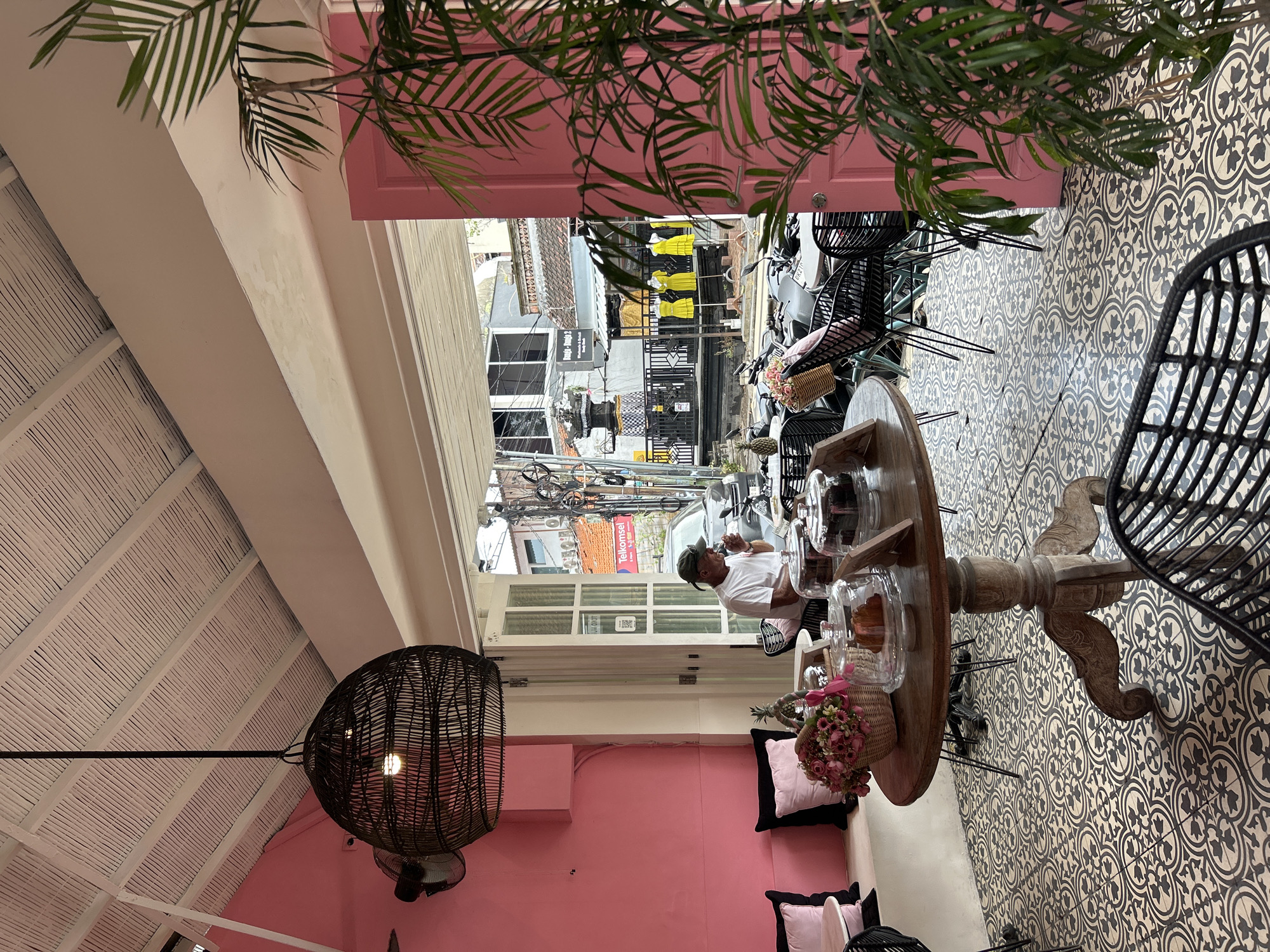 The image depicts the interior of a cafe in Kuta, Indonesia, with a focus on its vibrant pink walls and a patterned tiled floor. A man, wearing a green cap and a white t-shirt, is seated at a table, appearing to sip from a cup and eat something. The cafe features wicker furniture, including chairs and a large hanging lamp with a woven design. A round wooden table in the foreground holds a basket of flowers, pastries under glass domes, and a pineapple. The cafe opens onto a street scene visible through open double doors. Outside, there are motorcycles parked, a black car, and several mannequins displaying yellow dresses. The architecture visible outside includes traditional Balinese elements like a stone gate. The cafe's ceiling is white and lined with wooden planks. A large leafy plant is positioned in the foreground, partially obscuring the view of the street.
