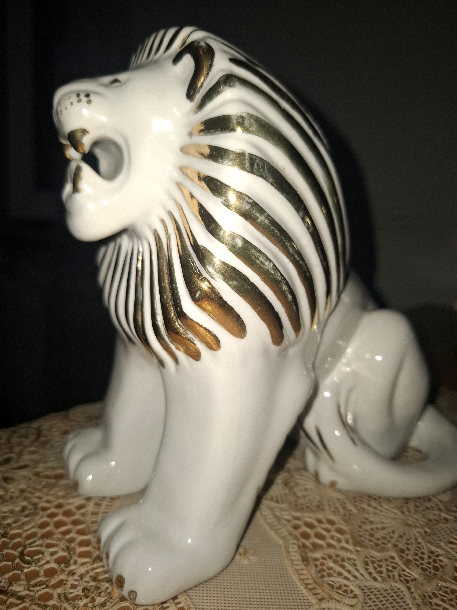 A white porcelain lion figurine with gold accents is positioned on a tan lace doily. The figurine is viewed from a side profile, with its head turned to the left. The lion has its mouth open, revealing its fangs. Its mane is depicted with alternating white and gold stripes. The doily has a honeycomb-like pattern and is placed on an unseen surface. The background is dark and out of focus. There is no indication of a specific location within the city of Almaty, Kazakhstan.