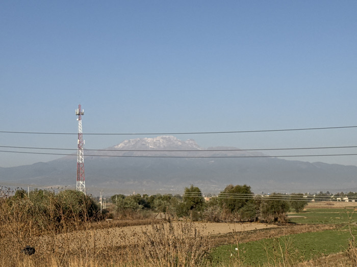 The scene depicts an expansive outdoor environment in Santa Ana Xalmimilulco, Mexico, under a clear, bright blue sky. In the immediate foreground, dry, light brown fields and overgrown, straw-colored vegetation dominate, partially obscuring the ground. Four dark, horizontal lines, consistent with utility wires, stretch across the middle third of the frame. To the left, a tall, lattice-structured cellular tower, painted in alternating red and white sections, rises prominently, featuring multiple antenna arrays near its apex. Beyond the foreground vegetation, segments of green cultivated fields are visible, interspersed with clusters of green trees and shrubs. In the distant midground, a low-density collection of light-colored, single-story structures suggests a settlement. The background is dominated by a large mountain range, its upper peaks partially covered with snow or ice, appearing somewhat hazy against the horizon. No people or animals are discernible, and no active interactions or specific actions are depicted within the static landscape. The overall lighting suggests daylight hours with direct sunlight.