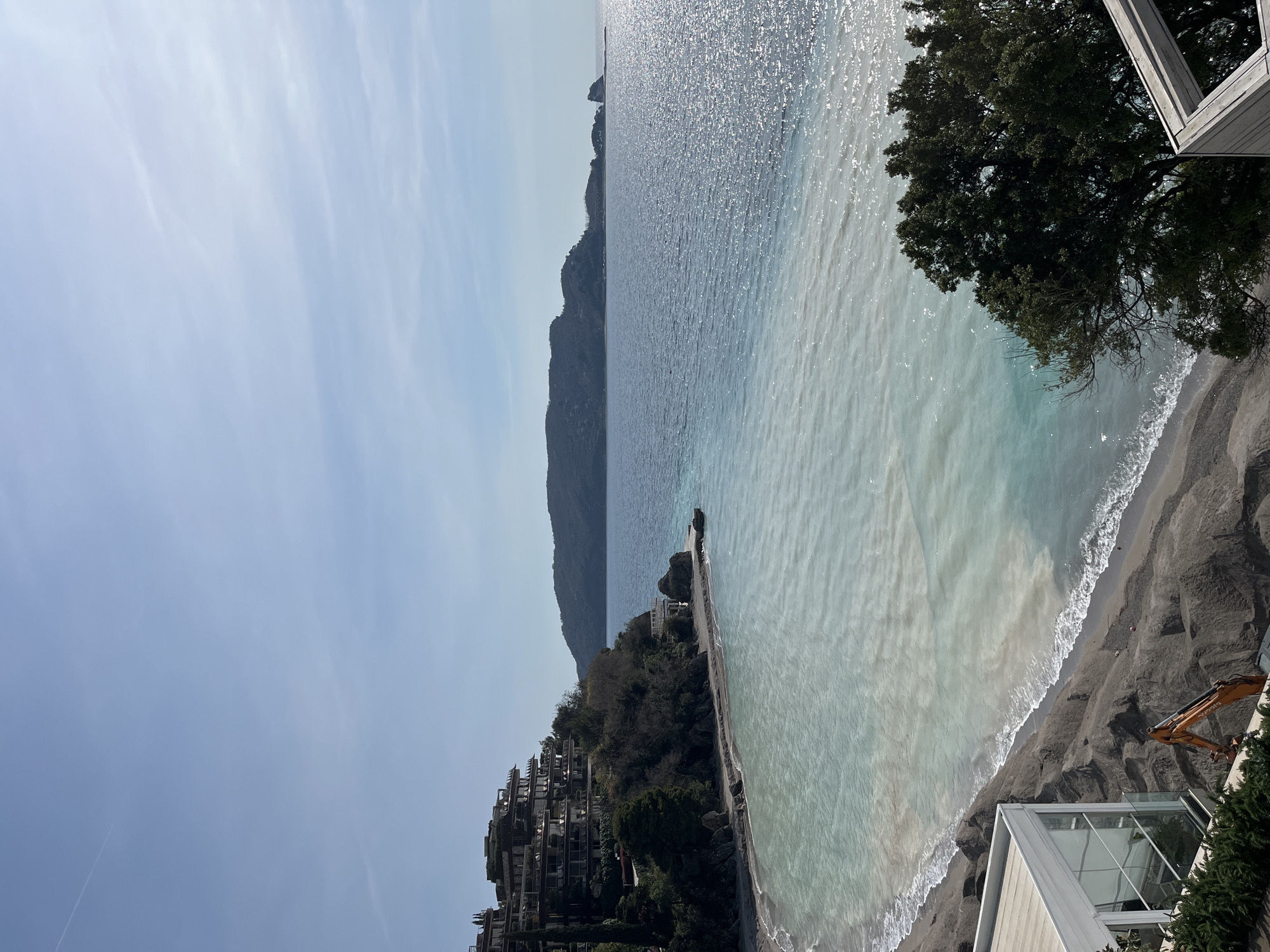 A wide-angle exterior view depicts a coastal scene in Budva, Montenegro, under a clear, light blue sky with a single faint contrail visible in the upper left quadrant. The image captures a bay with distinctly colored water. The nearshore water is an opaque, pale turquoise-to-grey, extending approximately one-third of the bay's width from the left shore before transitioning sharply into deeper, dark blue water that extends to the horizon on the right. Sun glare reflects off the surface of the darker water.
A dark grey, disturbed beach lines the left side of the bay, showing signs of recent earthworks. An orange excavator is partially visible in the lower-left corner, positioned on the disturbed earth near the water's edge. Directly above and to the left of the excavator, a modern building with a white frame and large glass panels is partially visible.
Further along the left coastline, a multi-story terraced building, possibly a hotel or residential complex, with numerous balconies and flat roofs, is integrated into a steep, verdant hillside. Dark green trees and shrubs surround and partially obscure this structure. A small, rocky outcrop extends into the water from the base of the hillside.
Across the bay, a prominent, dark green landmass with a rolling profile forms the distant horizon, accompanied by smaller rocky islets further to the right. No individuals are visible in the scene.