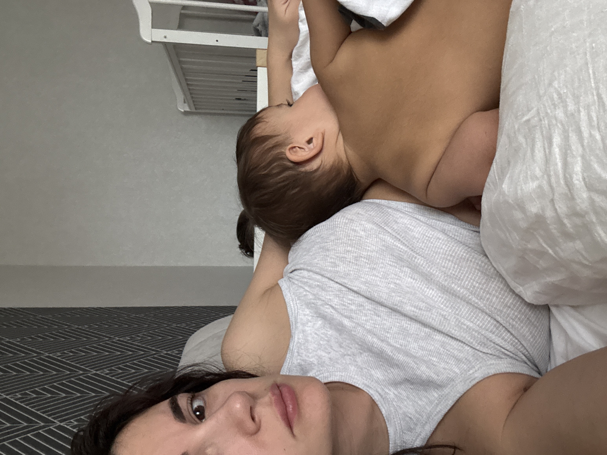 A woman is lying on a bed with a baby next to her, seemingly breastfeeding. The woman is wearing a gray tank top and her face is visible. The baby is partially covered by a white blanket. A white crib is partially visible in the background. The location is indoors. It is not possible to confirm that the location is in the city of Chaikovsky, Russia.