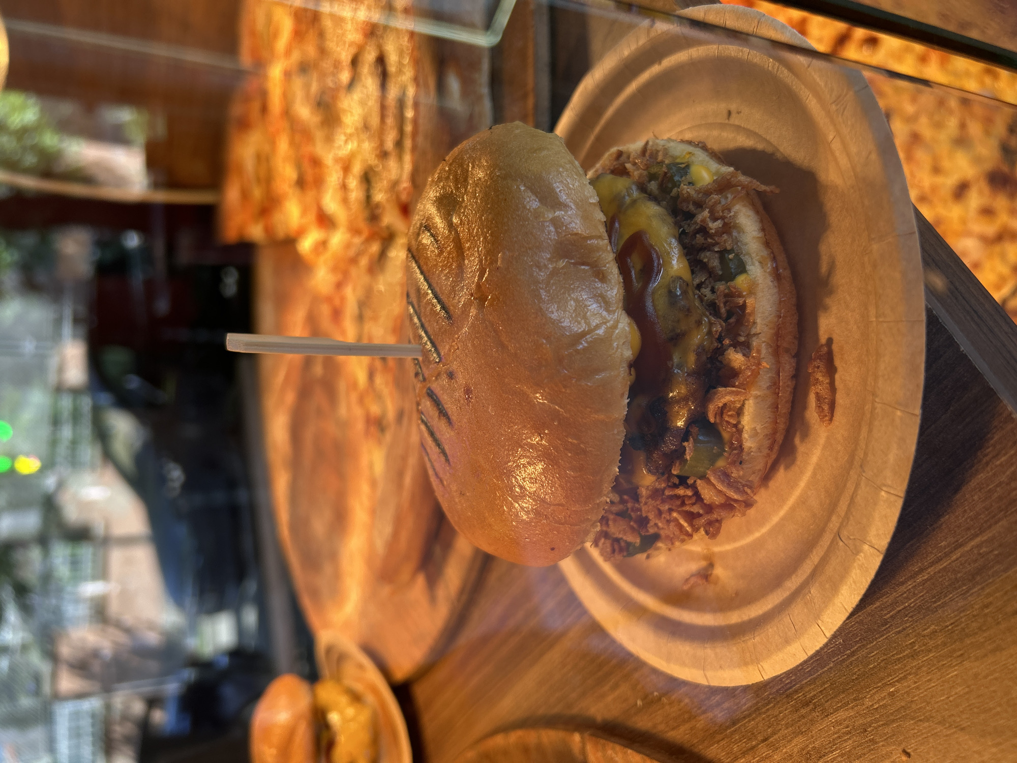 A burger sits on a paper plate, speared by a wooden pick. It is in the foreground, with pizzas in the background. The scene appears to be inside a restaurant or food establishment, possibly a fast-food place. The reflection in the glass shows a blurry image of a car and streetlights, suggesting an urban setting. The food is presented in a casual, takeout-style manner. The overall impression is of a quick meal, ready to be consumed. There are no people visible in the image. The setting is dimly lit, with the food being the main focus.