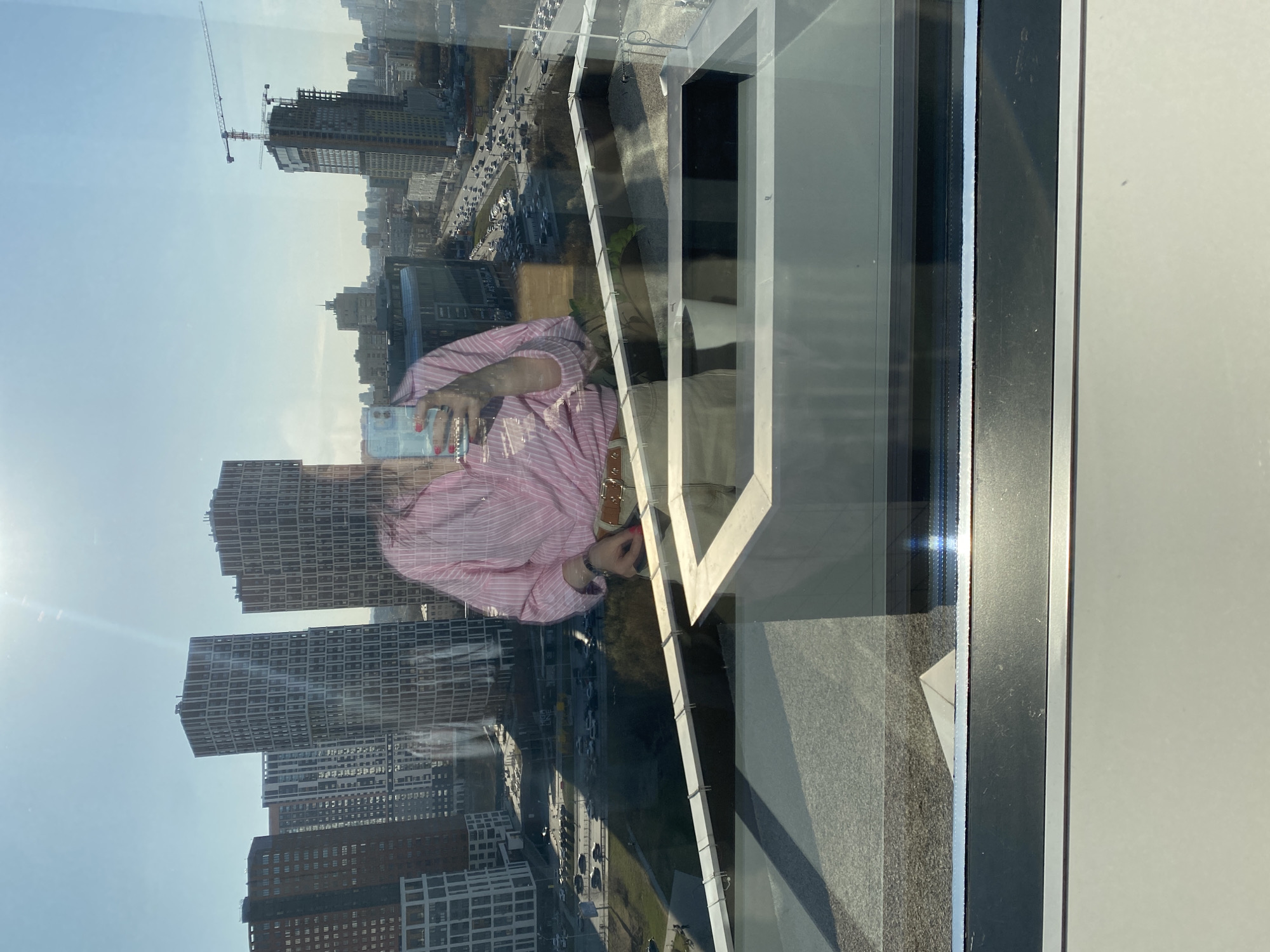 The image, when oriented correctly (rotated 90 degrees counter-clockwise), displays a high-angle view of an urban landscape in Yekaterinburg, Russia, captured through a window. The scene is bathed in bright daylight under a clear, sunny sky, with a notable sun glare and lens flare in the upper left corner.
In the foreground, a clear reflection of a person is visible, occupying the central-left portion of the frame. The person is wearing a pink and white striped long-sleeved shirt and appears to be taking a selfie with a smartphone, which they hold in their hand. Red nail polish is visible on their fingers. A light-colored handbag strap is also discernible over their shoulder.
Beyond the reflection, the cityscape unfolds. Numerous modern high-rise buildings with distinctive grid patterns cover the urban expanse. One prominent building is clearly under construction, featuring visible scaffolding and being serviced by a large red and white construction crane. Below these structures, a multi-lane road with cars can be seen, indicating a bustling urban environment. The clean lines of the window frame and a section of an outdoor balcony or architectural element are also present, adding depth to the composition. No legible text is visible in the image.