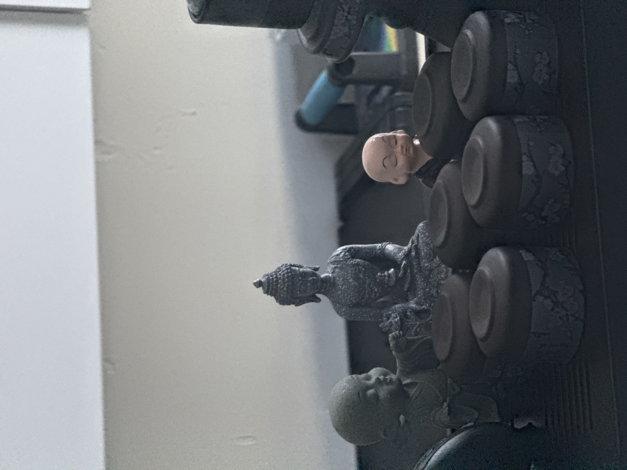 The image shows a close-up, eye-level view of several small decorative figurines and objects, likely on a shelf or table. In the center, a dark grey, intricately detailed Buddha statue is seated with its eyes closed. To the left of the Buddha, a small, stone-colored figurine of a young monk is partially visible, with one hand extended. To the right of the Buddha, a small, light-skinned figurine of a monk with closed eyes and a slight smile is positioned. In the foreground, arranged in a semi-circle, are at least four dark, lidded cylindrical containers. These containers appear to have a subtle floral pattern. The background is blurred, showing a light-colored wall and some indistinct objects, including what appears to be a blue handle of an implement. The overall scene suggests a collection of decorative items, possibly related to Buddhist or Asian culture. There is no indication of the city of Nha Trang, Vietnam, or any specific urban environment in the image.
