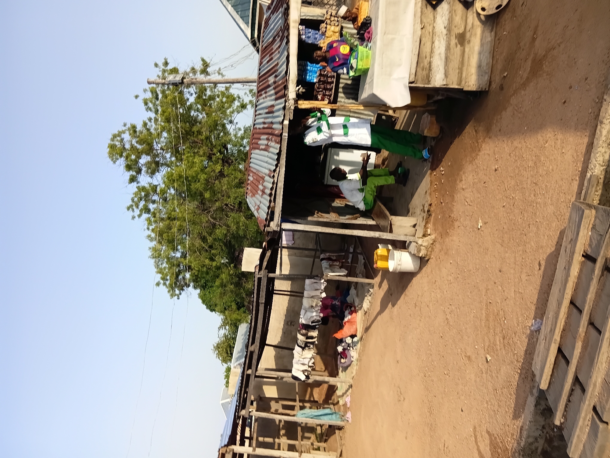 The image depicts an outdoor scene in Jalingo, Nigeria, characterized by a market stall and surrounding area. The stall, constructed with a corrugated metal roof, wooden posts, and siding, houses various items. A white refrigerator is visible inside the stall.
In front of the stall, several individuals are present. One person, wearing a white garment with green trim and green pants, stands with their arms crossed. Another individual, wearing a green tracksuit, is seated on a wooden bench. A woman, in the background behind a table covered with a white cloth, is looking down. The table displays goods, including a green basket filled with items and some packaged products.
Adjacent to the market stall, to the left, there is an open-air structure with a similar corrugated metal roof and wooden supports. Clothes are hanging on a line within this structure, and a pile of clothes is visible on the ground.
The ground is sandy and uneven. In the foreground, wooden planks are laid out. Several buckets and a yellow container are also visible on the ground. Trees are present in the background, and electrical wires are strung overhead, indicating the presence of infrastructure. The sky is clear and pale blue, suggesting daytime.