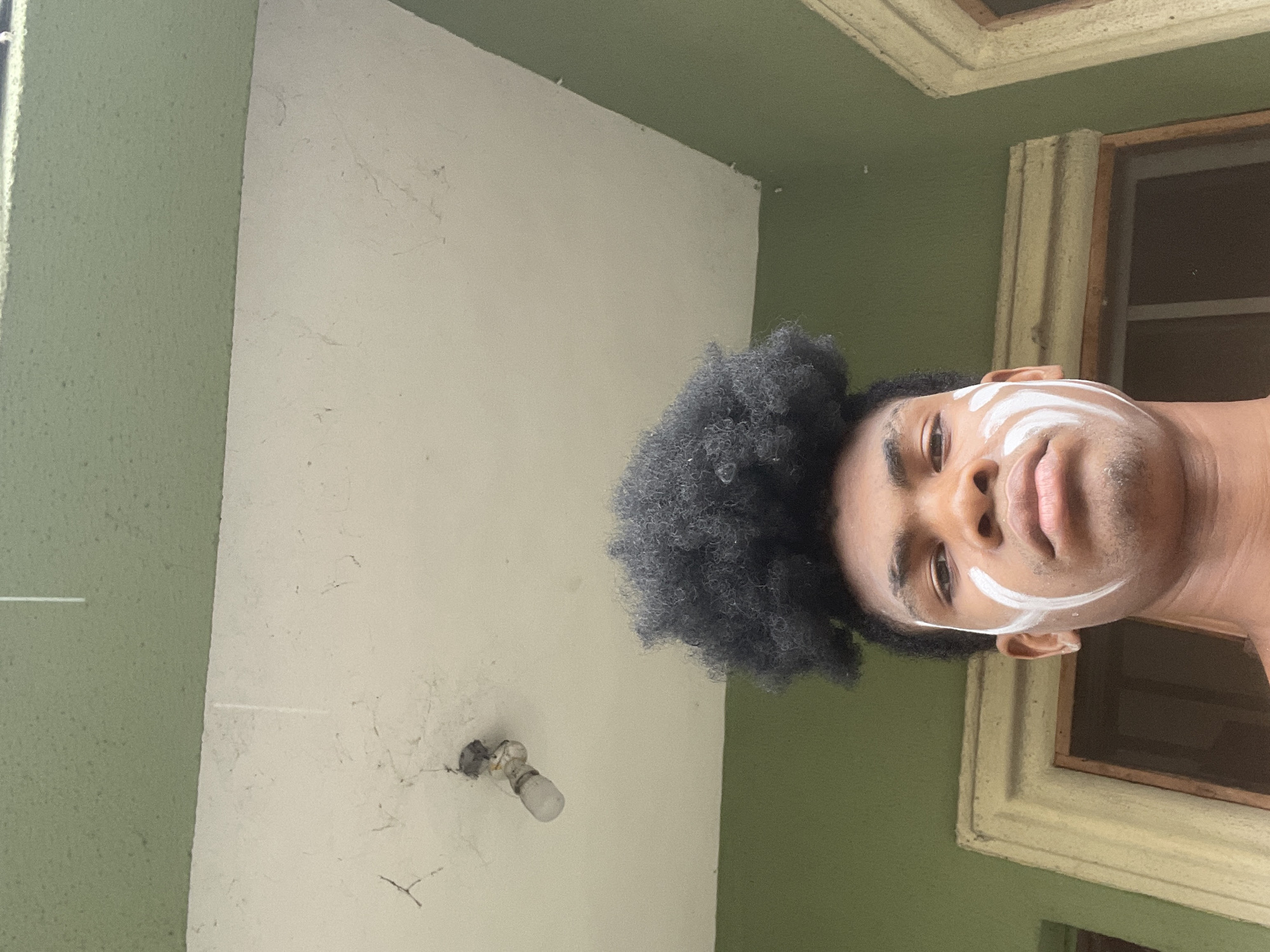 A low-angle shot shows a man's face with a dark afro hairstyle against a green and white indoor background. He has white paint-like markings on his face, appearing as a curved line near each ear and another along the upper cheek. A window with beige frames and vertical bars is visible to his right. The wall is divided into green and white sections, and a bare light bulb fixture is on the white portion. The location is likely Ado, Nigeria, but there are no location-specific details to confirm within the image itself.