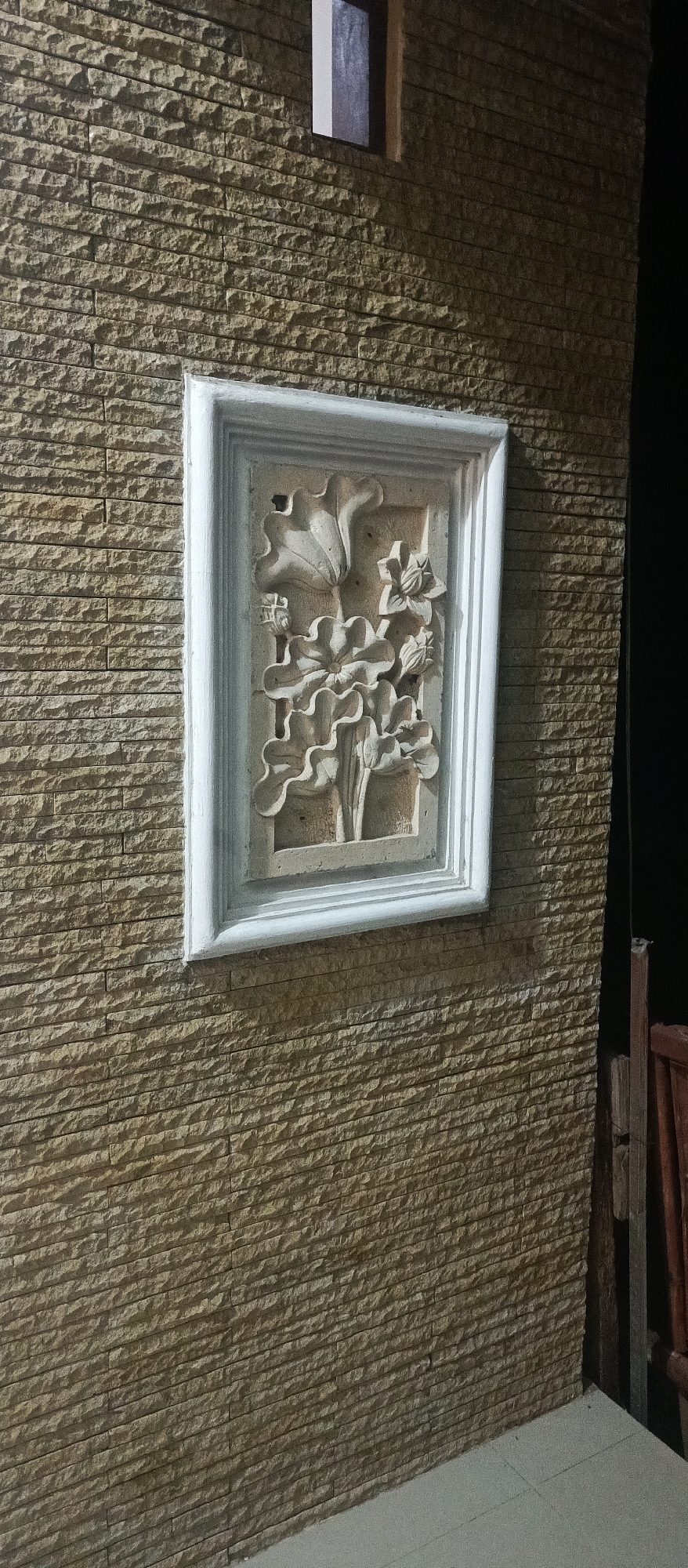 The image shows a close-up, eye-level view of a textured stone wall with a decorative relief panel. The wall is constructed of horizontally stacked, rectangular stones in varying shades of beige and brown, creating a rough, uneven surface. The relief panel, framed by a white, beveled border, is mounted centrally on the wall. This panel features a sculpted design of lotus flowers and leaves, rendered in a lighter, sandy-colored material. The relief is detailed, with distinct petal shapes and vein patterns on the leaves. Above and to the left of the relief, a small, dark rectangular opening, possibly a window or vent, is visible against the stone wall. To the right of the relief, a dark, indistinct area suggests a shadow or an adjacent structure. The lighting appears to be artificial and cast from above, creating subtle shadows that highlight the texture of the stone and the depth of the relief. The lower portion of the image shows a light-colored tiled floor meeting the base of the wall at an angle. The scene appears to be an exterior wall, potentially part of a building or garden feature, at night.