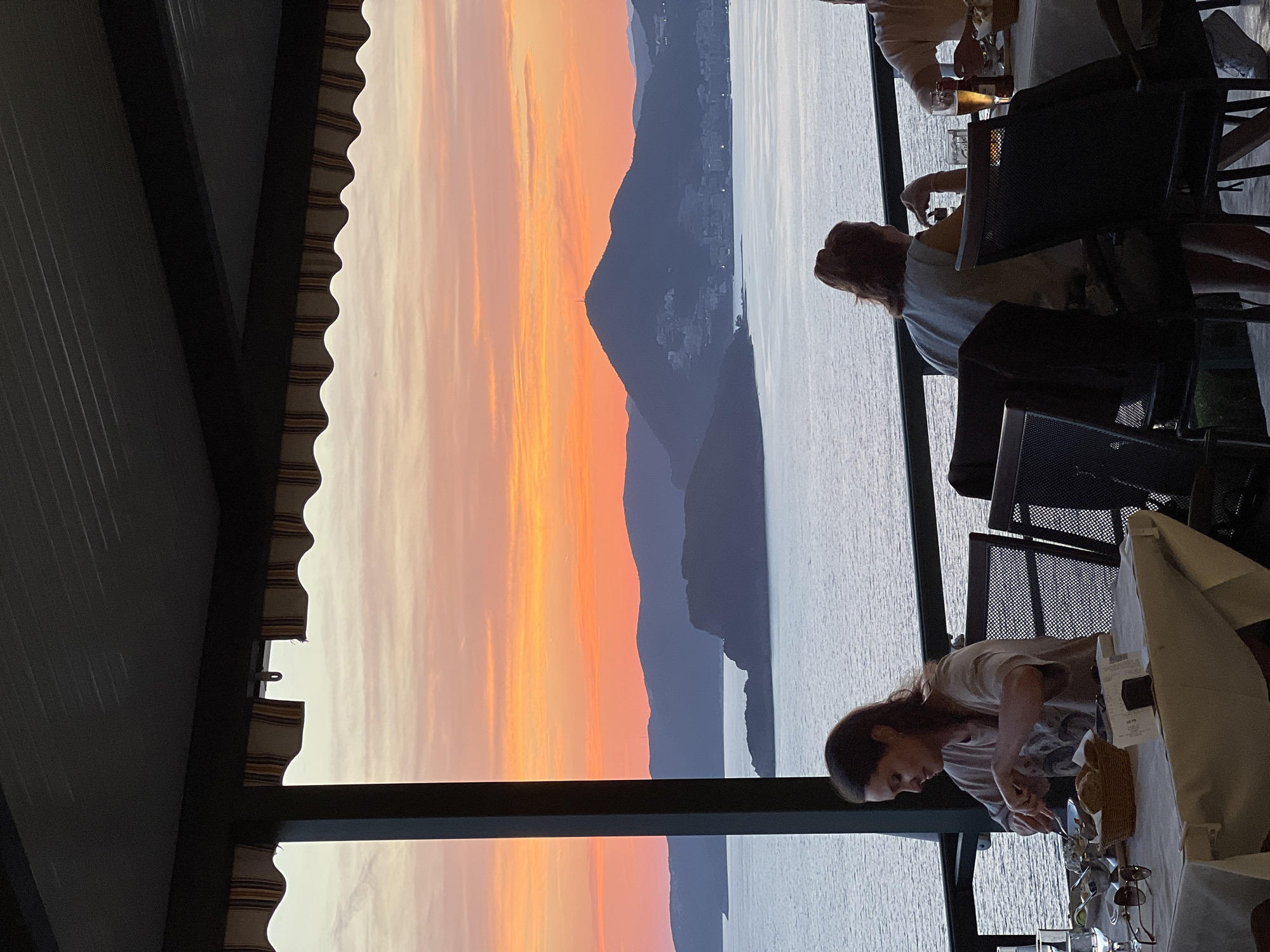 The image shows a partially obstructed sunset view from a restaurant in Sveti Stefan, Montenegro. A partial awning is on the left side of the frame. A large body of water stretches to the horizon, separating the restaurant view from the silhouette of distant mountains.
There are people at tables in the foreground. Two are seated and two are standing at the tables. One person appears to be cutting food, while another has their back to the camera and is seated. Various dining objects are visible, including plates, glasses, utensils, and chairs.
