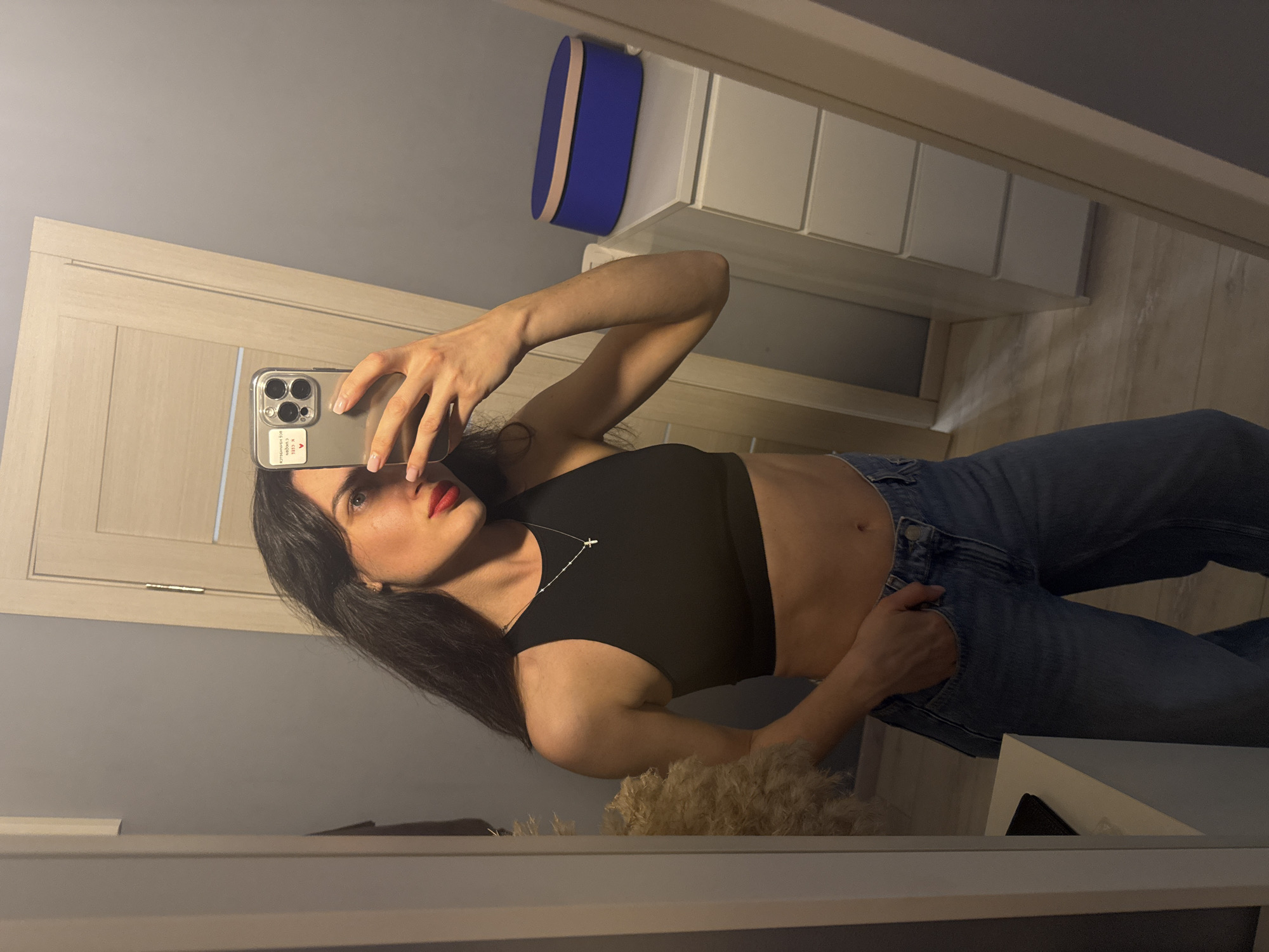 A mirror reflects a woman taking a selfie with an iPhone in what appears to be a room. The woman has long, dark hair and wears a black crop top and jeans. Her left hand is in her pocket. A door is visible in the background, and some storage units are stacked with a blue box on top. The location appears to be indoors, possibly a bedroom, but the specific city and broader location are unknown.