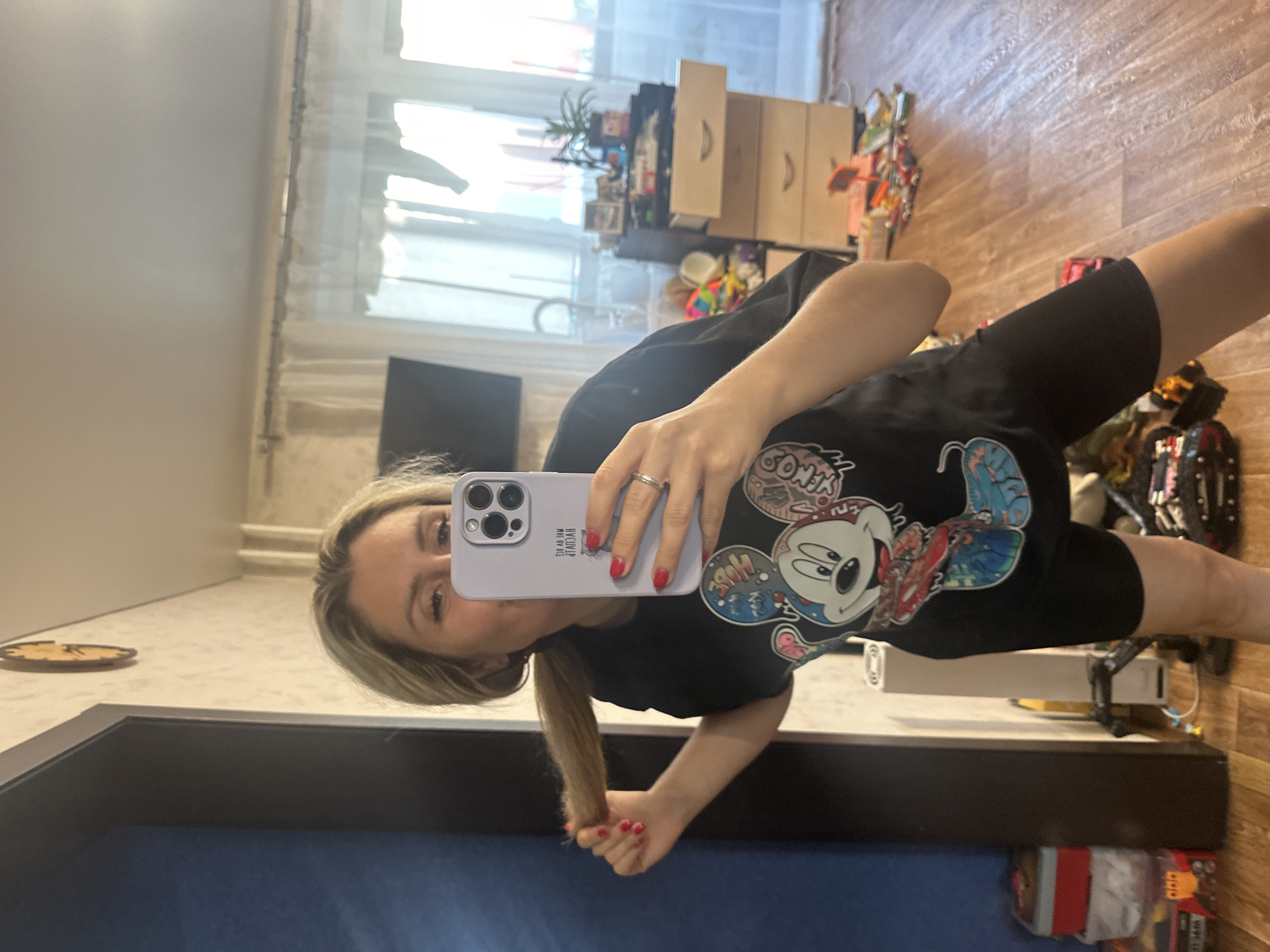 This is a vertically oriented, medium-shot, self-portrait taken in a room, likely a bedroom or living area. The primary subject is a young woman with blonde hair, wearing a black t-shirt with a graphic print of Mickey Mouse and other cartoon characters. Her nails are painted red. She is holding a light purple smartphone in her right hand, which is reflecting her face and upper body in a mirror. The background shows a wooden floor, a window with blinds, and several pieces of furniture including a dresser with drawers and shelves filled with various items. A plant is visible on a shelf. The room appears to be indoors, and the lighting suggests it could be daytime, possibly late afternoon given the quality of light filtering through the window. The woman's expression is difficult to determine precisely due to the angle and reflection, but she appears to be posing for the photo. Some text is visible on the back of her t-shirt, but it is too small and distorted to be legible. The overall impression is a casual, personal snapshot.