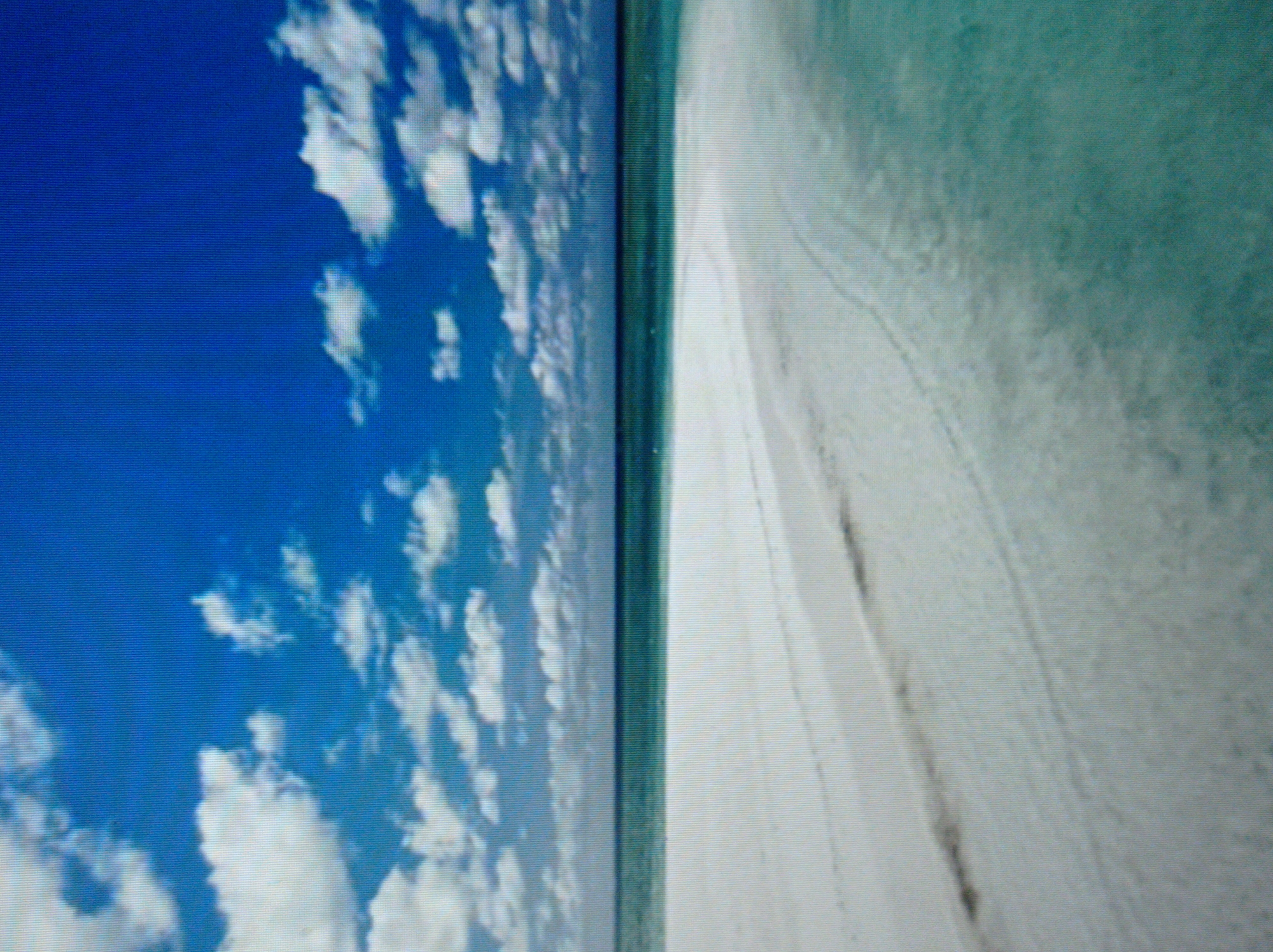 The image depicts a wide, sandy beach with the turquoise ocean stretching to the horizon under a bright blue sky filled with scattered white clouds. The sand appears smooth with subtle ripples and some darker streaks, possibly from wet sand or debris. The water is clear and shallow near the shore, gradually deepening to a darker hue further out. There are no people or man-made objects visible in the scene, suggesting a remote or natural setting. This scene is not located in the city of Mukachevo, Ukraine, as it depicts a coastal environment, and Mukachevo is an inland city.