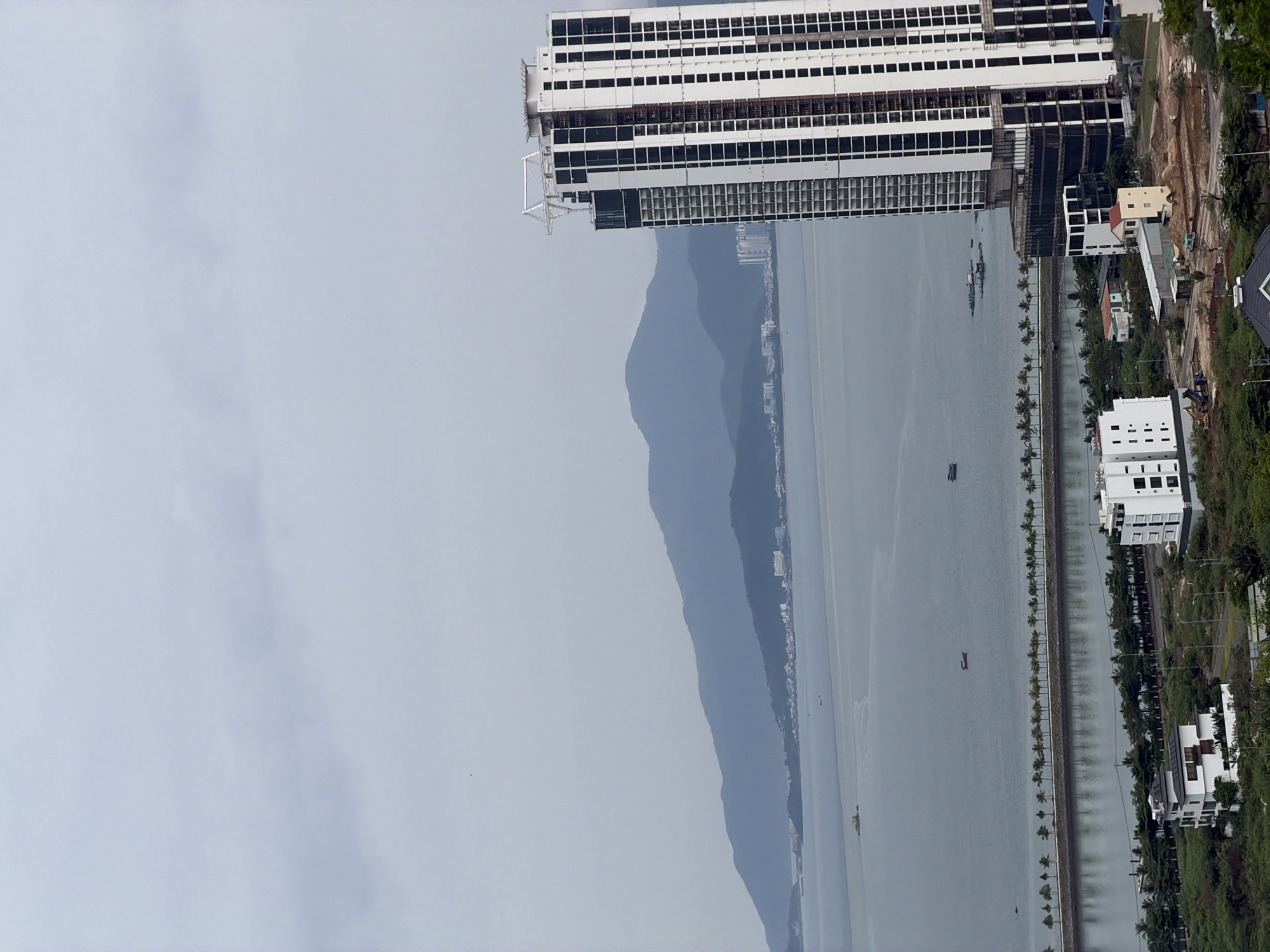 View of Bac Nha Trang Ward, Nha Trang, Vietnam. A multi-story, unfinished white building stands parallel to the top edge of the frame, next to a body of water with boats. Across the water are two mountains. Several buildings line the water's edge to the right with palm trees.