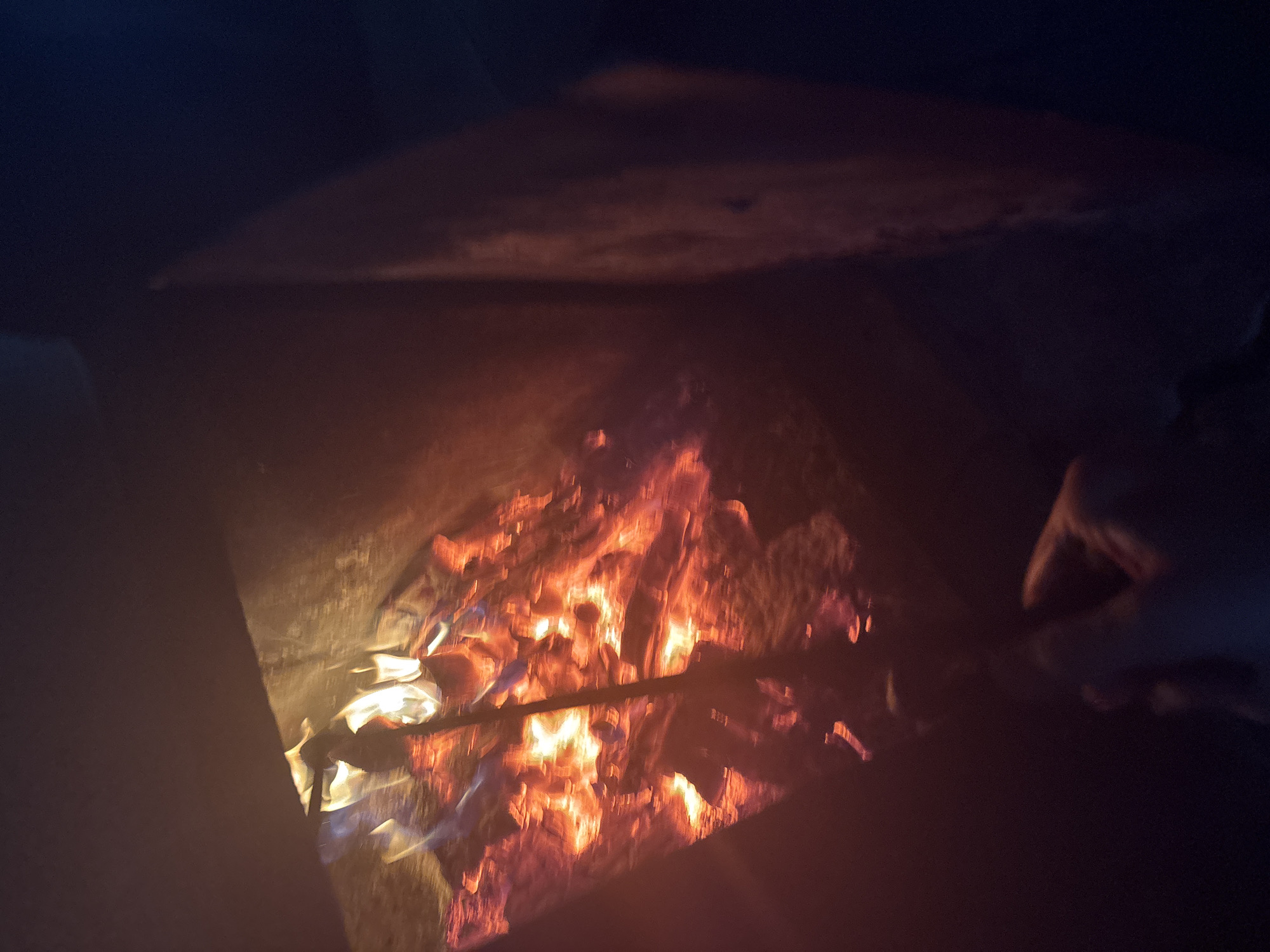 The image shows a close-up view of a fire burning in what appears to be a makeshift fire pit or brazier. The flames are a vibrant orange and yellow, casting a warm glow on the surrounding surfaces. The fire is contained within a dark, metallic structure, the sides of which are visible. A hand, partially obscured, is holding a dark-colored rod or skewer, seemingly inserted into the fire. The background is dark, with some indistinct shapes that could be part of the environment or objects. The overall impression is one of a person tending a fire, possibly for cooking or warmth. There is no specific indication of location within the city of Курган, Russia.