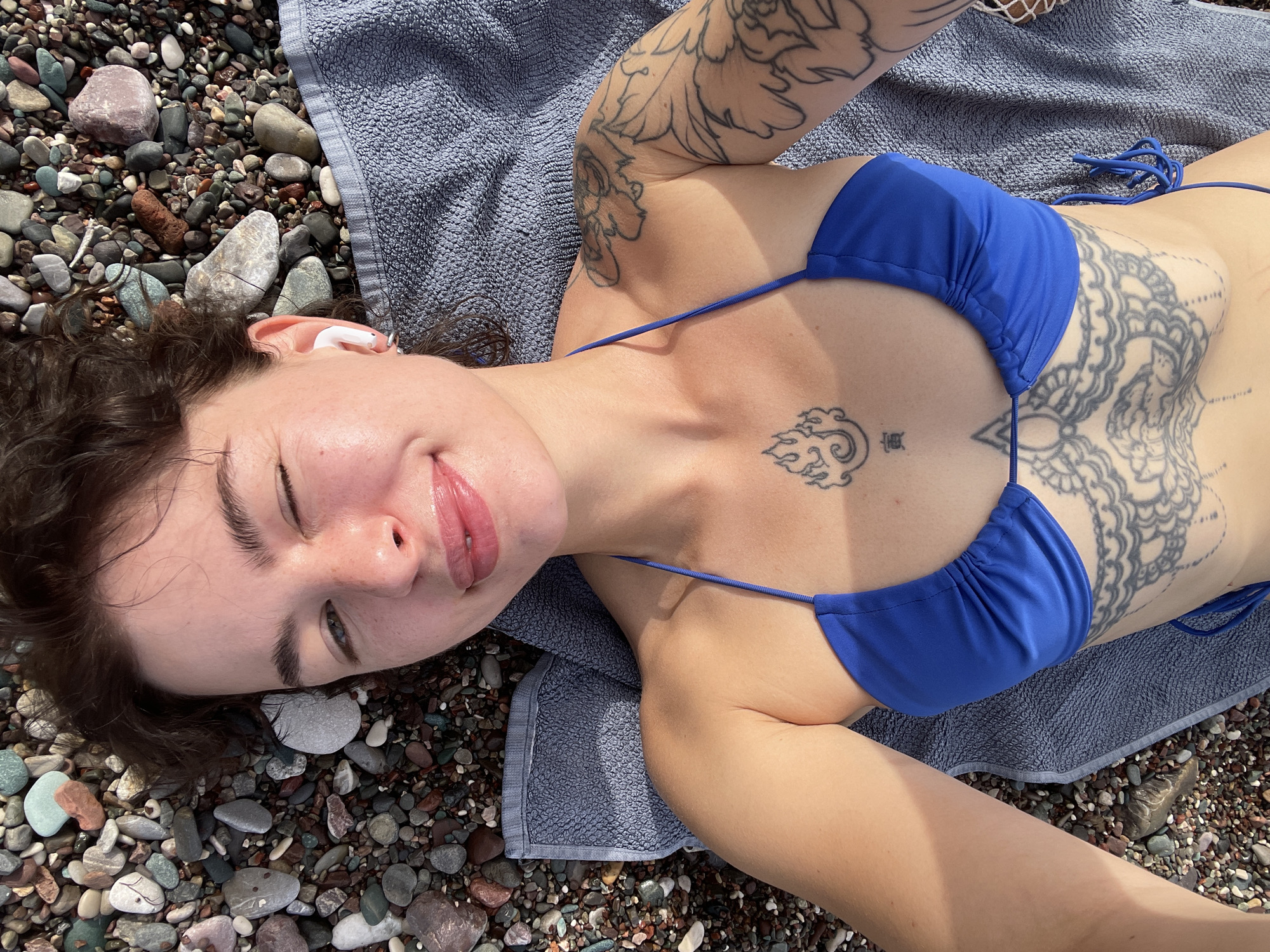 A young woman is captured in a high-angle shot, lying on her back on a rocky beach in Rafailovići, Montenegro. She is wearing a blue bikini top and has dark hair, with an earbud visible in her right ear. Her face is directed towards the camera, featuring a relaxed smile and a playful wink of her right eye, conveying a happy and carefree mood.
She is positioned on a grey towel, which is laid out over a bed of diverse, multi-colored pebbles and rocks that form the beach's surface. The bright, direct sunlight suggests it is daytime, likely mid-day or afternoon, indicating clear weather.
Several tattoos are prominent on her body. A large, intricate floral and leafy design covers her right arm (left side of the image). On her chest, below her collarbone, is a smaller tattoo featuring a stylized cloud or wave pattern, with the Chinese character "壽" (shòu), meaning longevity, inked beneath it. Additionally, a large, detailed mandala-like tattoo is visible on her right torso and rib cage, partially obscured by her bikini top. The scene depicts a moment of relaxation and enjoyment in a sunny, natural beach environment.