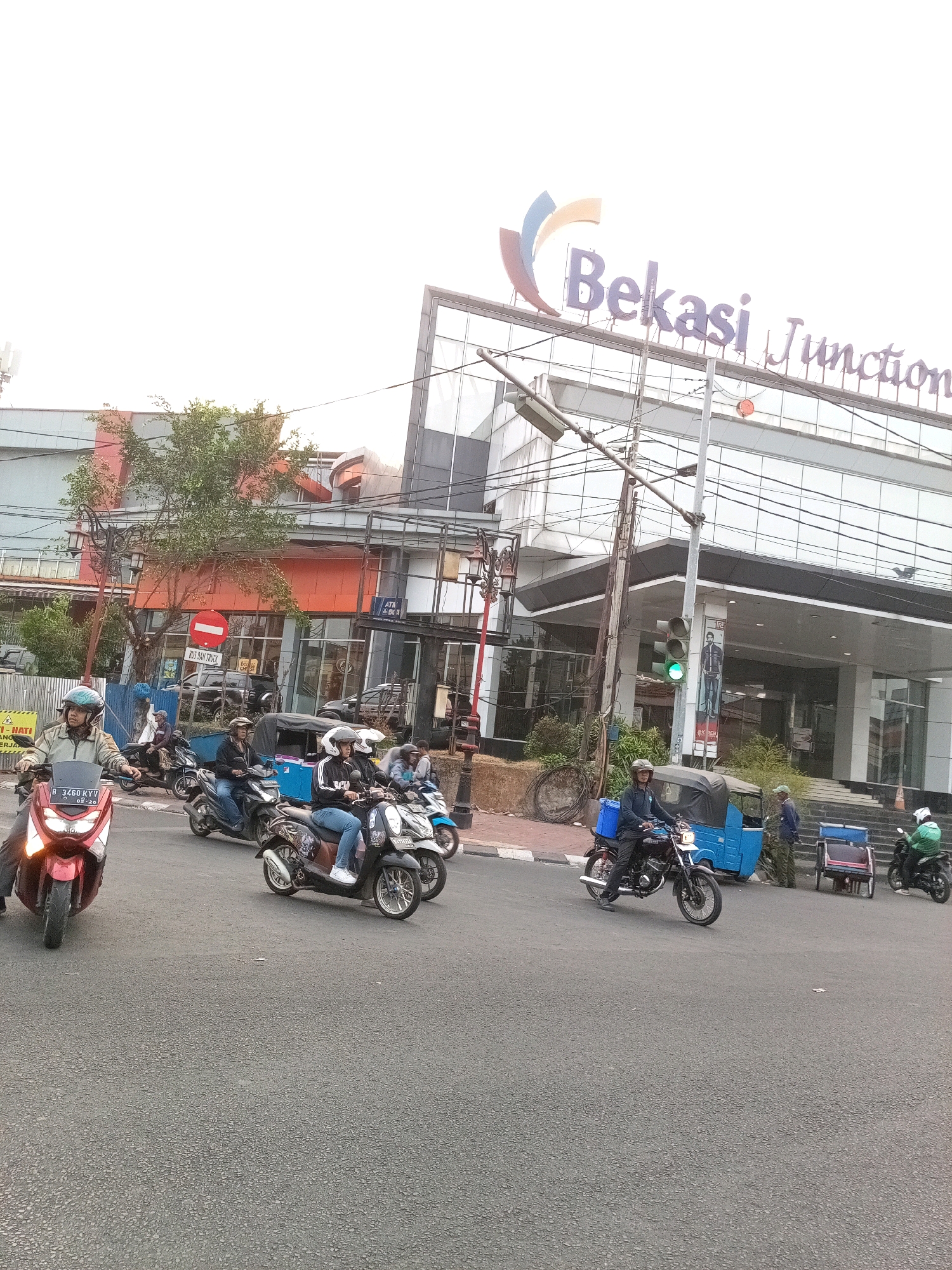 This is an eye-level, medium shot of a street scene in Bekasi, Indonesia, featuring traffic and a commercial building. In the foreground, several motorcycles are visible on the asphalt road. One red scooter is in the left foreground, with its front wheel turned slightly. Behind it, a group of people on motorcycles are moving across the street. A blue three-wheeled vehicle is also present. In the background, the modern, multi-story "Bekasi Junction" building dominates the scene, with its glass facade reflecting the sky and signage displaying the name of the establishment. Several trees and utility poles are also part of the urban landscape. The overall atmosphere suggests a busy commercial district.