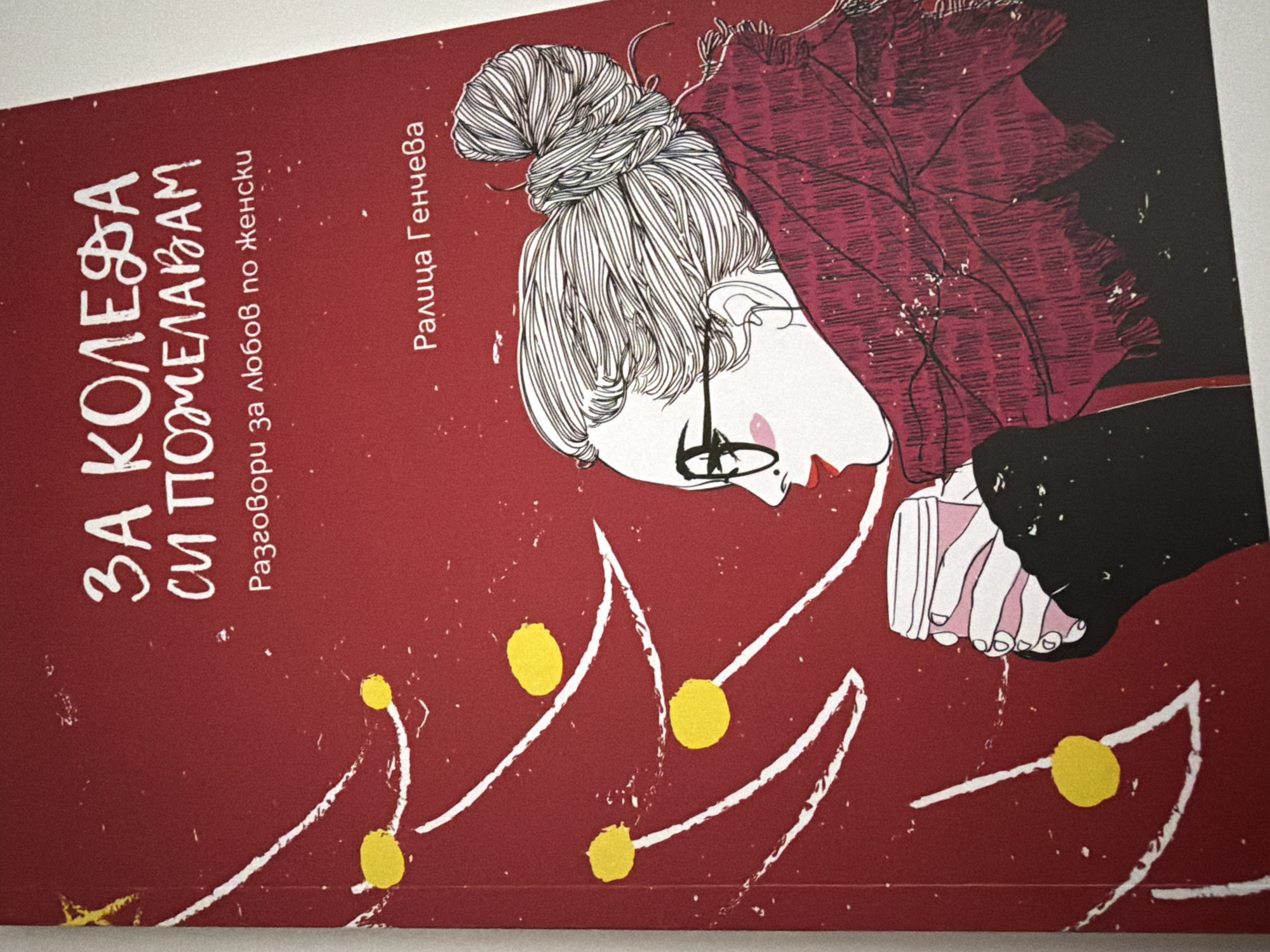 The image is a book cover with a red background and a Christmas theme. The title, written in white, says "ЗА КОЛЕDA СИ ПОЖЕЛАВАМ" which translates to "For Christmas I Wish You." Below this, in smaller white text, it says "Разговори за любов по женски" which translates to "Conversations about love, women's style." At the bottom right, the author's name is written in white: "Ралица Генчева".
The illustration on the cover features a stylized Christmas tree on the left, with white branches and yellow ornaments. On the right side, there is a drawing of a woman from the profile, facing left. She has her hair in a bun and is wearing glasses and a large, textured red scarf. She is holding a pink and white disposable coffee cup in her hands.
There is no information in the image to indicate a specific location in the city of Pazardzhik, Bulgaria.