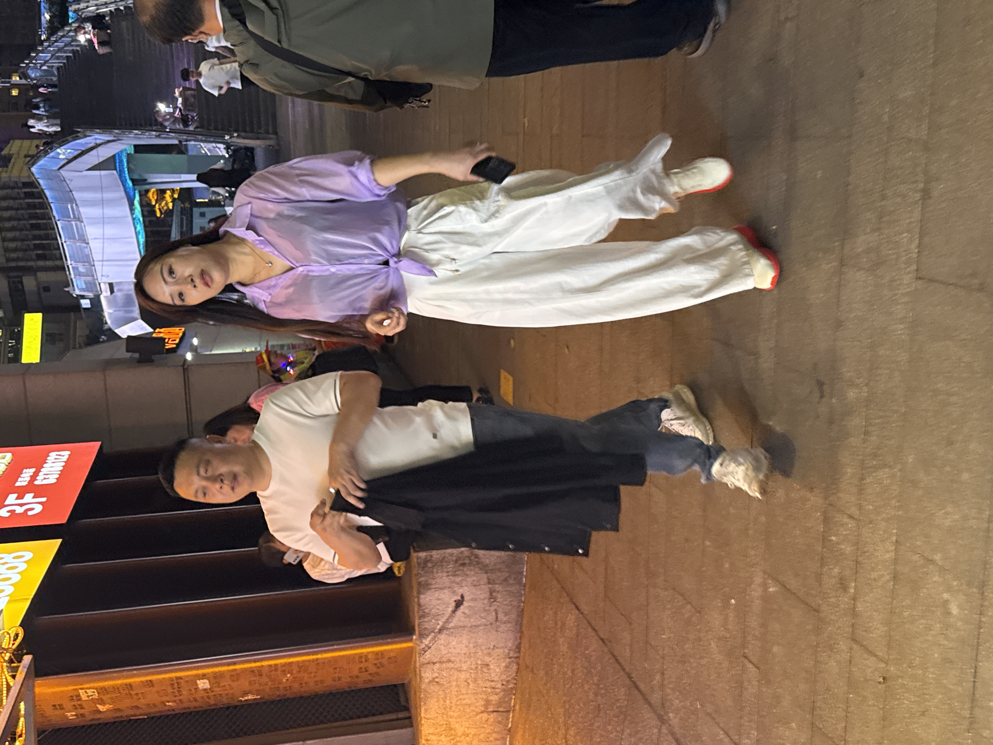 In Yuzhong District, China, a woman in a purple shirt and white pants lies on the sidewalk holding a phone. Nearby, a man in a white shirt and jeans holds a black jacket. The scene is illuminated by street lights and building signs. In the background are escalators and a covered walkway.