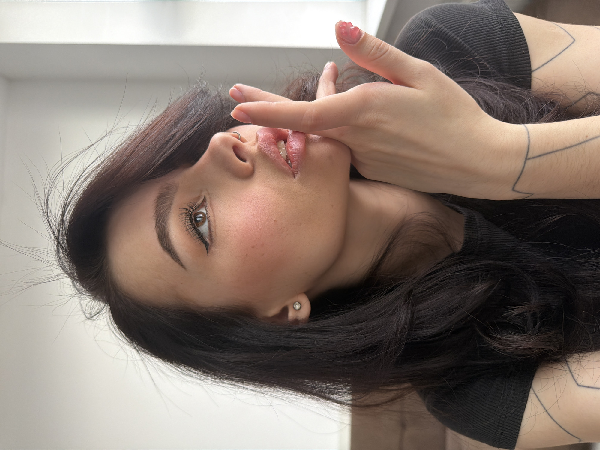 The image is a close-up portrait of a person, rotated 90 degrees clockwise. The subject is a person, likely female, with long, dark hair that frames her face and flows downwards. She has fair skin, light brown eyes, and dark, well-defined eyebrows. Her head is tilted slightly, and her eyes are open, looking upwards and to the right of the frame.
One of her hands is raised, with fingers partially covering the lower part of her face, specifically her chin and mouth. Her lips are slightly parted, revealing a glimpse of her teeth. The nails on her visible hand are manicured and coated with a translucent pink or clear gel polish, which appears thick and somewhat textured, especially on the thumb.
Notable facial features include a prominent cat-eye eyeliner, long eyelashes, and light blush on her cheeks. She wears a small silver hoop nose ring on her left nostril and a small stud earring in her right earlobe. She is wearing a black, ribbed top or shirt. Geometric line tattoos are visible on her left arm and shoulder area.
The background is a simple, light, and unfocused area, suggesting an indoor setting with soft, natural lighting, likely during the daytime. There is no visible text in the image. The overall expression conveyed by the subject is thoughtful or enigmatic.