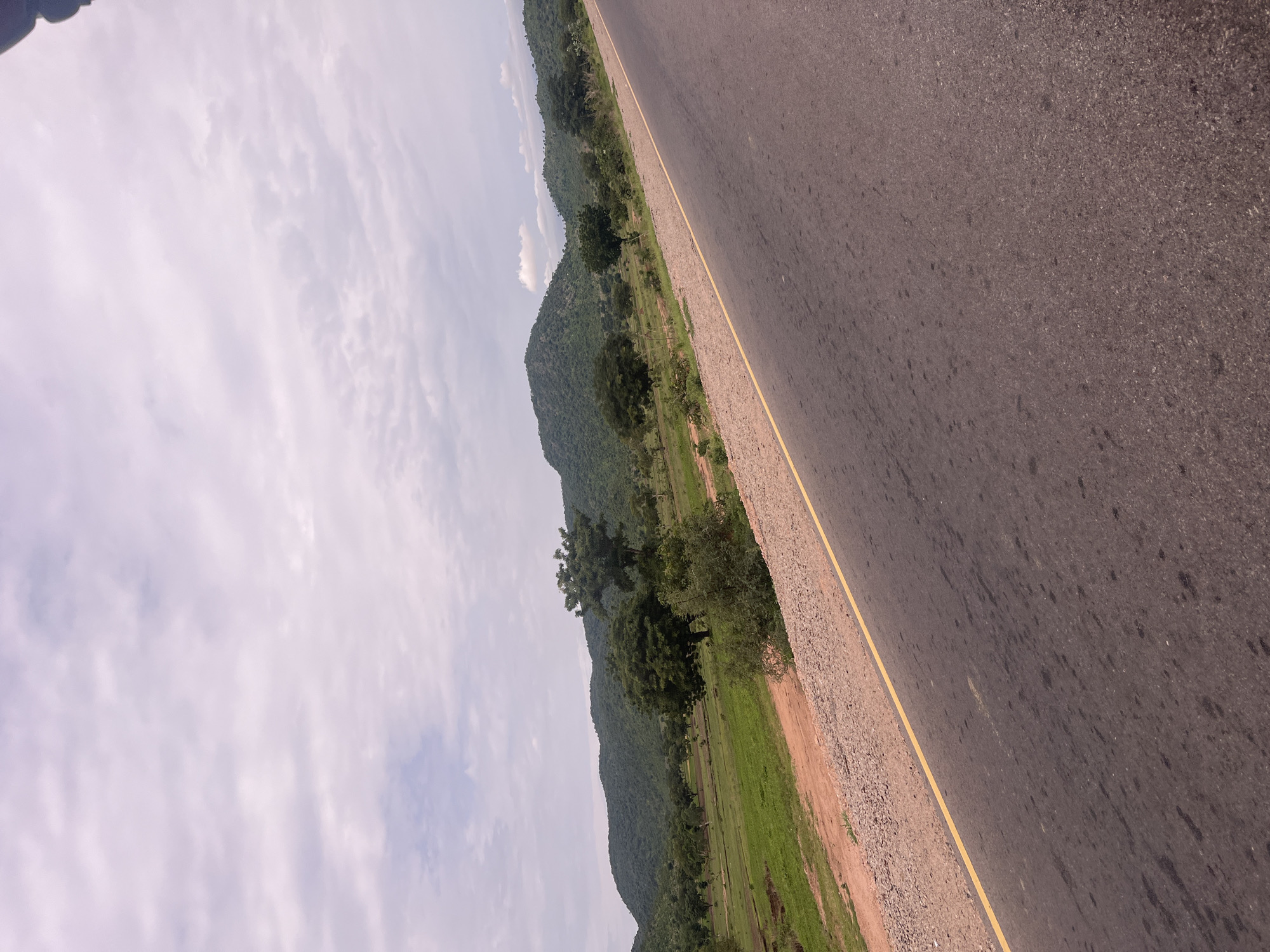 This media file is a landscape photo, taken from the perspective of someone traveling on a paved road. The road, with a yellow line marking its edge, occupies the right side of the frame and extends diagonally into the distance. To the left of the road, there's a strip of gravelly earth, followed by a vibrant green field that stretches towards a backdrop of lush, tree-covered hills. The hills are undulating and covered in dense vegetation, indicating a natural, possibly rural or undeveloped area. The sky above is partly cloudy, with streaks of white and grey clouds against a lighter sky, suggesting daytime. There are no people or specific objects visible, creating a sense of solitude and focus on the natural scenery. The overall impression is one of travel through a scenic, possibly tropical or subtropical, environment. The location context provided is Minda, Nigeria, which aligns with the visual cues of green hills and vegetation. The image appears to be taken during daylight hours, with no specific time of day being evident beyond that. The weather seems fair, with clouds present but not obscuring the sun. There is no visible text in the image.