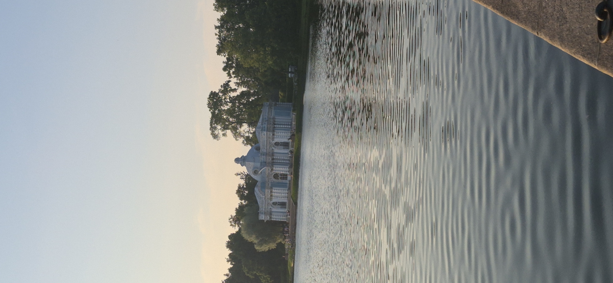 The image depicts a tranquil scene with a pavilion situated near a body of water. The pavilion, with its ornate architecture, features blue and white detailing and domes, suggesting a classical or Baroque style. It is surrounded by lush green trees, hinting at a park or garden setting. The water, with its gentle ripples, reflects the soft light of the sky, which is a pale blue, transitioning to a warmer hue near the horizon, indicating either sunrise or sunset.
The image's orientation is vertical, suggesting it might be a portrait-style photograph. The focus is divided between the architectural structure and the shimmering water. The water occupies the right half of the image and exhibits a textured surface due to the wave patterns. A stone ledge, possibly part of a dock or pathway, is visible in the upper right corner, adding a textural contrast to the water.
There are a few indistinct figures near the pavilion, suggesting the presence of people, but they are too small to discern any specific activity or emotion. The overall mood is peaceful and serene, enhanced by the soft lighting and the natural elements. The absence of harsh shadows and the calm water suggest fair weather. The specific location is not discernible from the image. No text is visible in the frame.