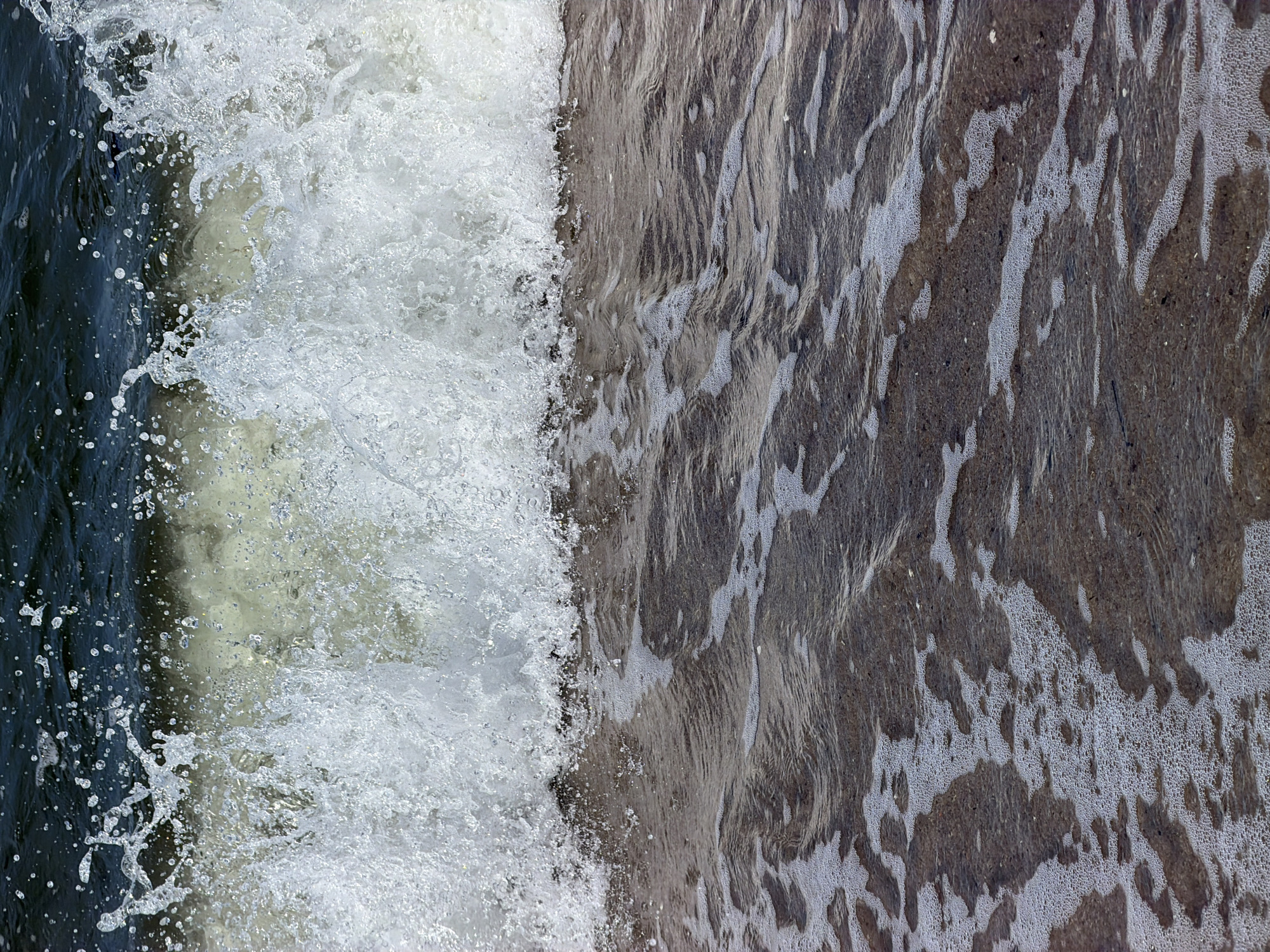 A close-up, eye-level shot captures a wave crashing onto a dark, sandy shore. The wave is in mid-break, with a crest of white foam and spray dominating the upper portion of the image. The water itself is a deep, dark blue, with lighter, foamy elements visible beneath the crest. The shore below is composed of dark sand with small pebbles, and is covered in a thin layer of receding foamy water, creating intricate patterns. The scene is set in Baltiysk, Russia.