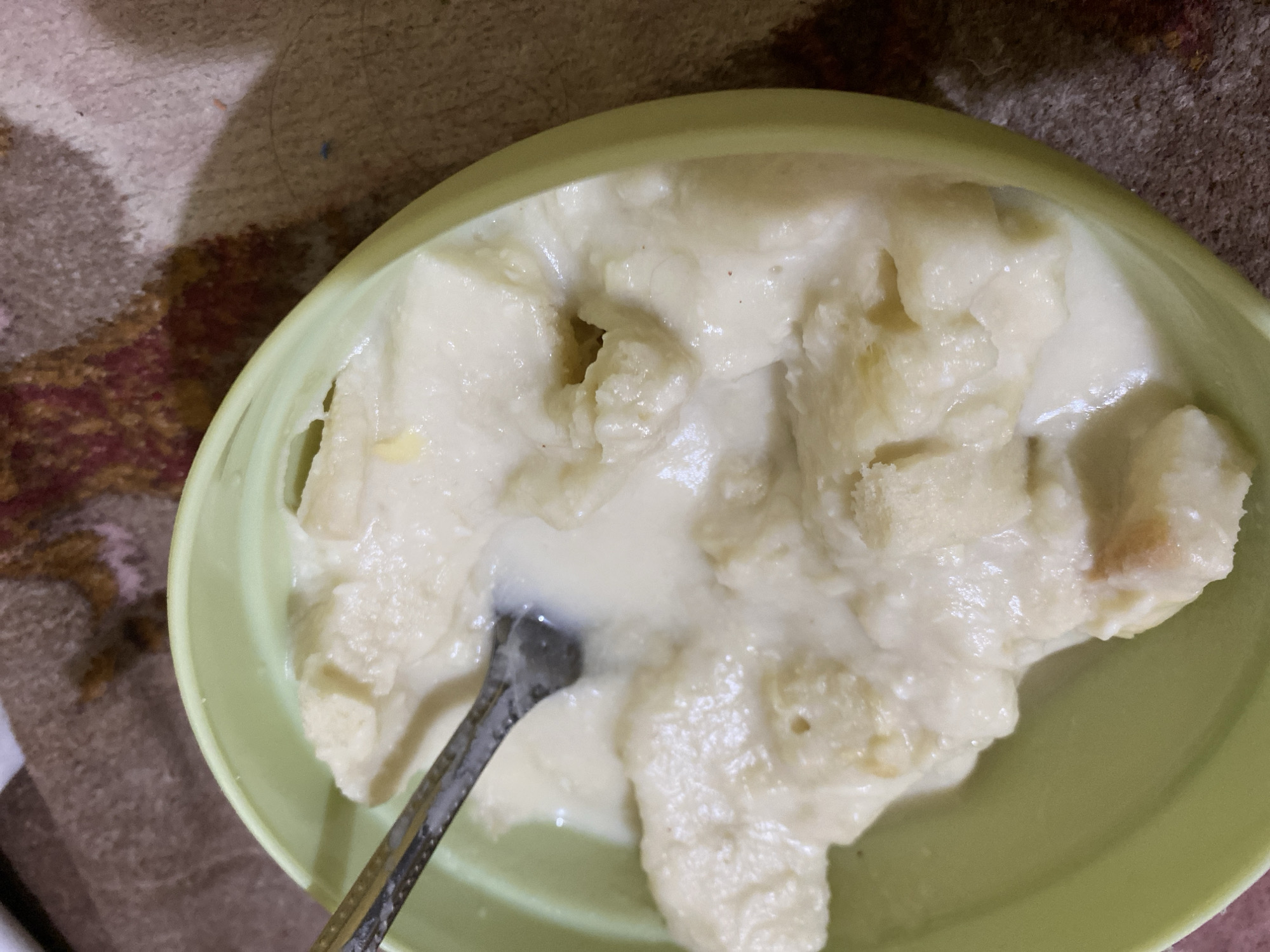 A bowl of bread pudding with a fork is shown in a close-up shot. The pudding is made of soft white bread chunks mixed in a creamy, white sauce. A silver fork is partially submerged in the pudding, suggesting it is being eaten. The bowl is light green and sits on a surface with a brown, patterned texture, possibly a tablecloth or blanket. The lighting is somewhat dim, creating soft shadows. The scene appears to be indoors, in a domestic setting, likely in Lambaro Kueh, Indonesia, where bread pudding is a common dessert.