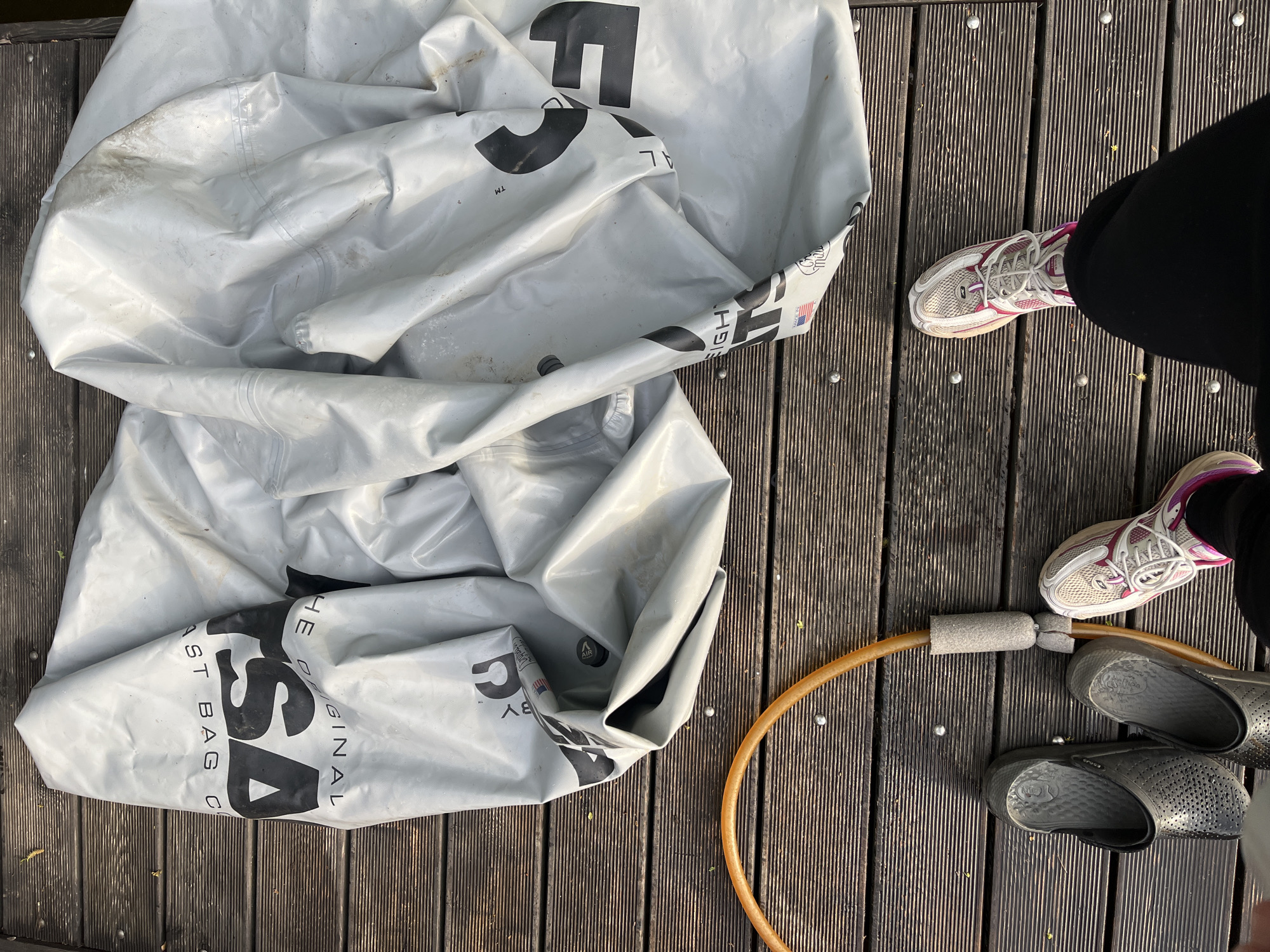 The image shows a section of a weathered wooden deck. On the left side, a large, deflated, light gray vinyl bag with the words "THE ORIGINAL LAST BAG CO." printed in black is crumpled. To the right of the bag, a person is standing with their feet visible. They are wearing running shoes (white and pink) and black slip-on shoes (gray). Between the shoes is a section of orange hula hoop that connects with a gray foam cushion. The location is not possible to know.