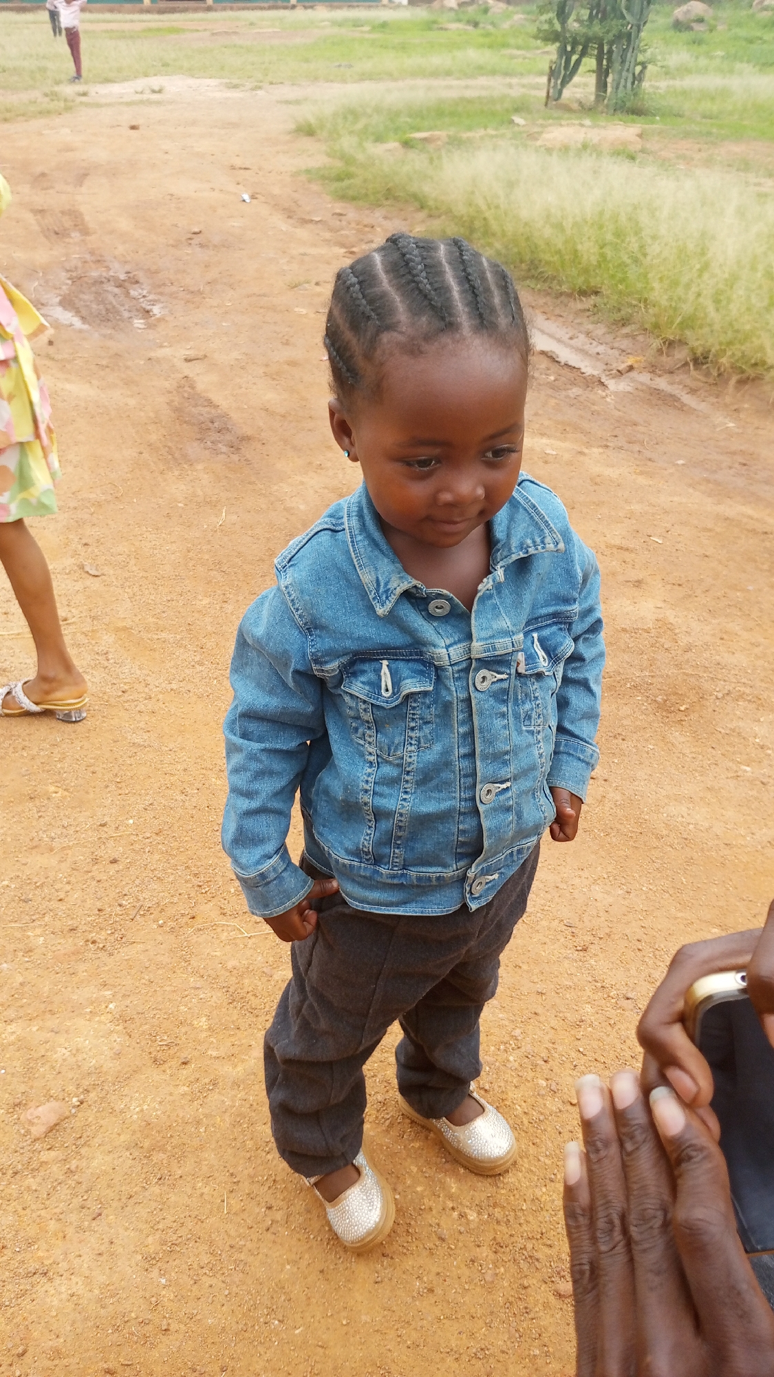 A young child with braided hair stands on a dirt surface in what appears to be an outdoor environment, possibly near Barikin Sabon Gidan Kanat, Nigeria. The child wears a denim jacket, dark trousers, and light-colored shoes. Another person's legs and feet are visible on the left side of the frame, while a hand holding a smartphone is on the right. The background features sparse vegetation and a building in the distance.
