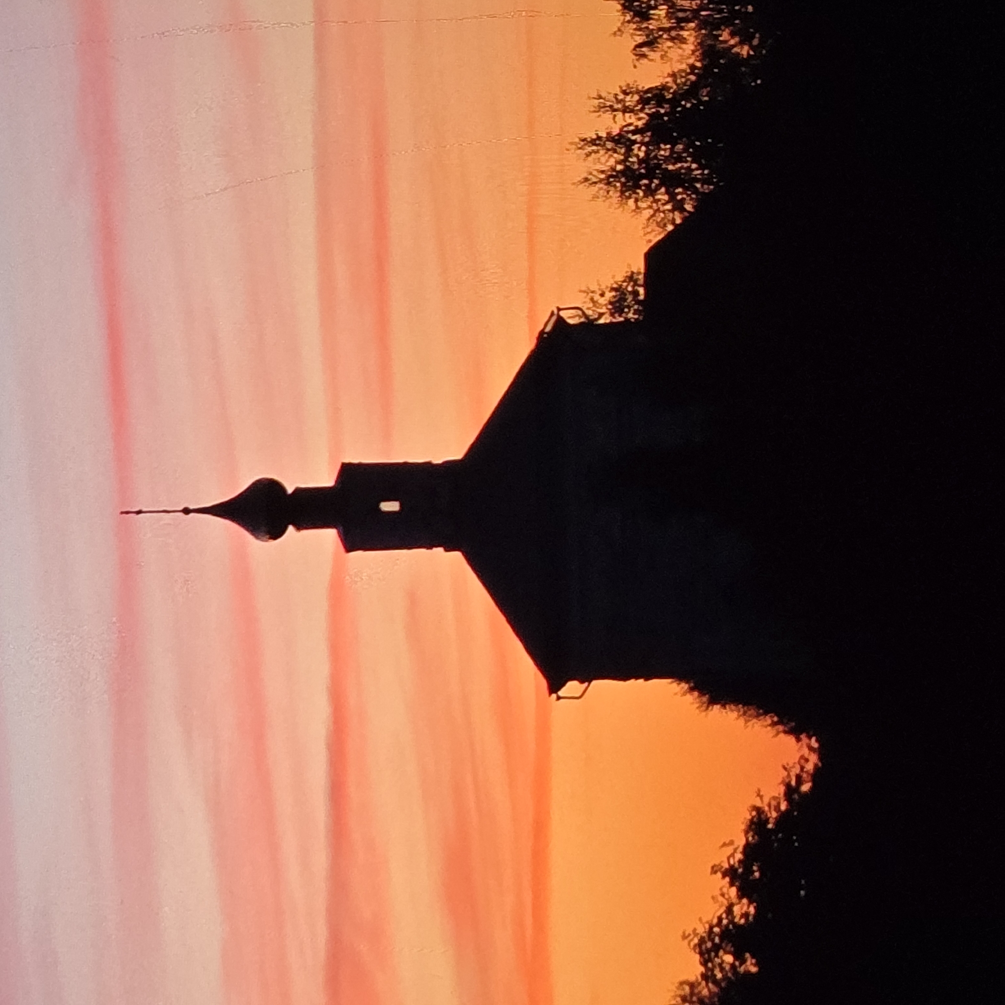 The image shows a silhouetted church against a vibrant sunset sky. The sky transitions from a deep orange at the bottom to pink and lighter shades of orange at the top, with horizontal cloud streaks. The church has a distinctive onion dome topped with a spire and a small cross. There is a rectangular opening visible in the upper portion of the church's structure. In the foreground and along the bottom of the image, the dark silhouettes of trees frame the scene. The overall impression is one of a peaceful, rural setting during twilight. The church is likely part of a rural settlement or a historically significant site in the Vladimir Oblast, Russia, given the architectural style common in the region.
