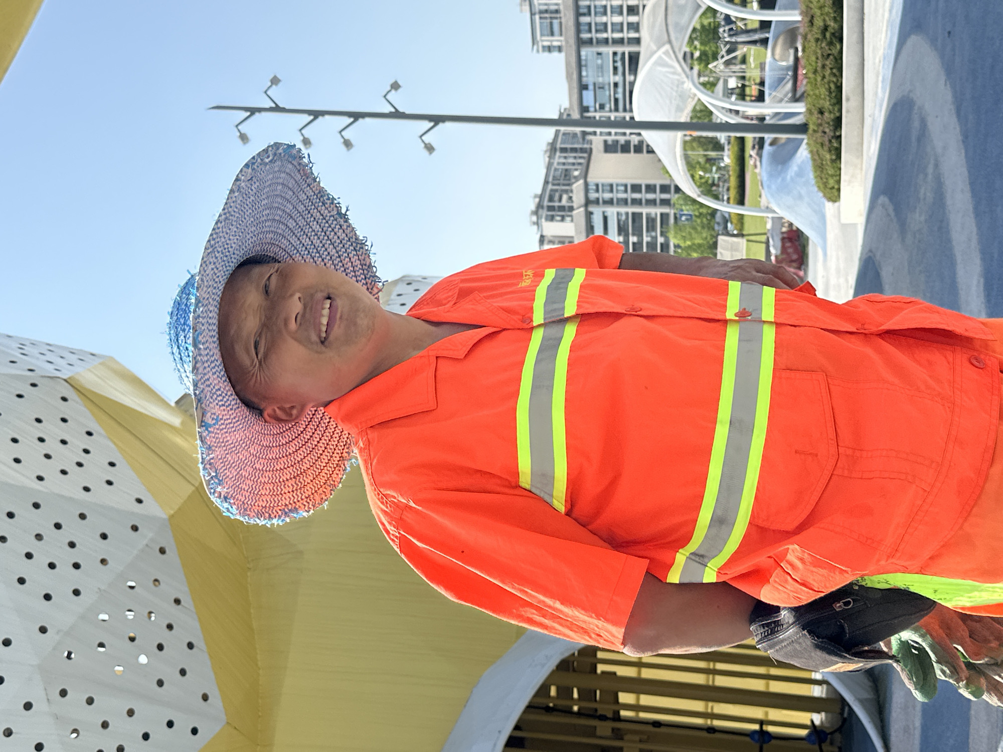 An outdoor portrait, taken on a bright, sunny day, features a smiling man. The image is oriented with the sky on the left and the ground on the right, but when viewed correctly, the man stands facing slightly upwards and towards the viewer with a broad, cheerful smile, revealing his teeth. He appears to be of East Asian descent, likely middle-aged or older.
The man is wearing a light blue and white woven straw hat that shades his head. His attire includes a vibrant orange short-sleeved high-visibility work shirt, accented with prominent vertical reflective stripes in yellow and grey across the chest. On the left chest area of his shirt, Chinese characters are visible, which read "环卫" (Huánwèi), indicating his role as a sanitation worker. He also appears to be wearing orange pants, though only a small section is visible. A black baseball cap and a pair of green work gloves are tucked into his belt or waistband on his right side.
The background suggests a modern urban park or public space, consistent with a location like Jiangjin, China. Under a clear blue sky, several tall, contemporary buildings with reflective glass facades are visible in the distance. Distinctive architectural elements include a large, white, curvilinear structure and a yellow, geometric structure with perforated panels, possibly part of a contemporary art installation or play area. The ground features sections of blue-colored pavement and some green shrubbery. The bright sunlight and lack of clouds indicate a clear weather day, likely mid-morning or afternoon. The man's happy expression conveys a positive mood.
