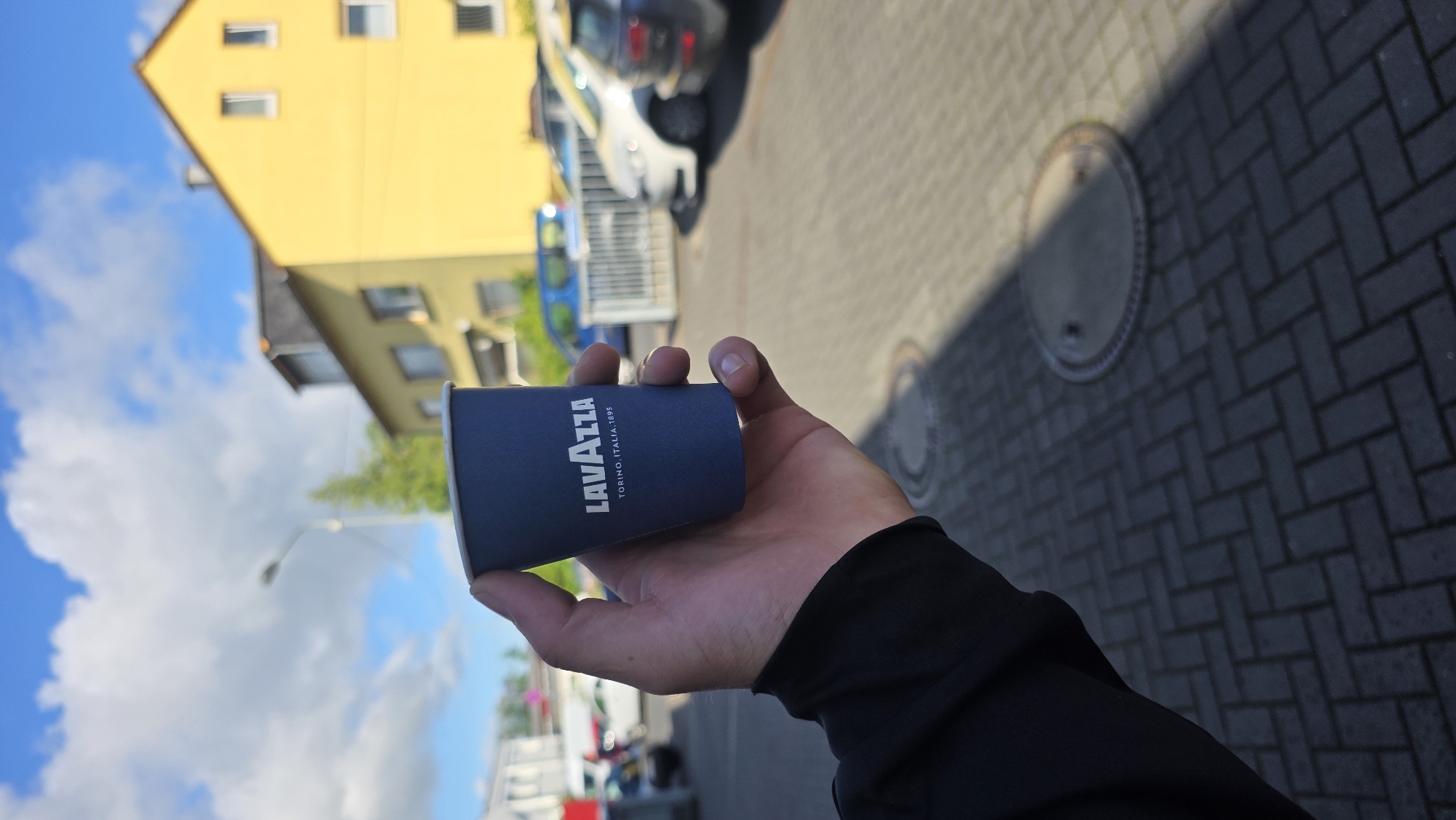A hand holds a small, dark blue Lavazza coffee cup against a backdrop of a yellow building and a blue sky with clouds. The cup has the Lavazza logo and "TORINO, ITALIA, 1895" printed in white. The hand is holding the cup from the side, and the person's arm is visible, wearing a black jacket. In the background, there are parked cars, a street light, and a paved ground with a brick pattern. The scene appears to be in an outdoor urban or suburban setting.