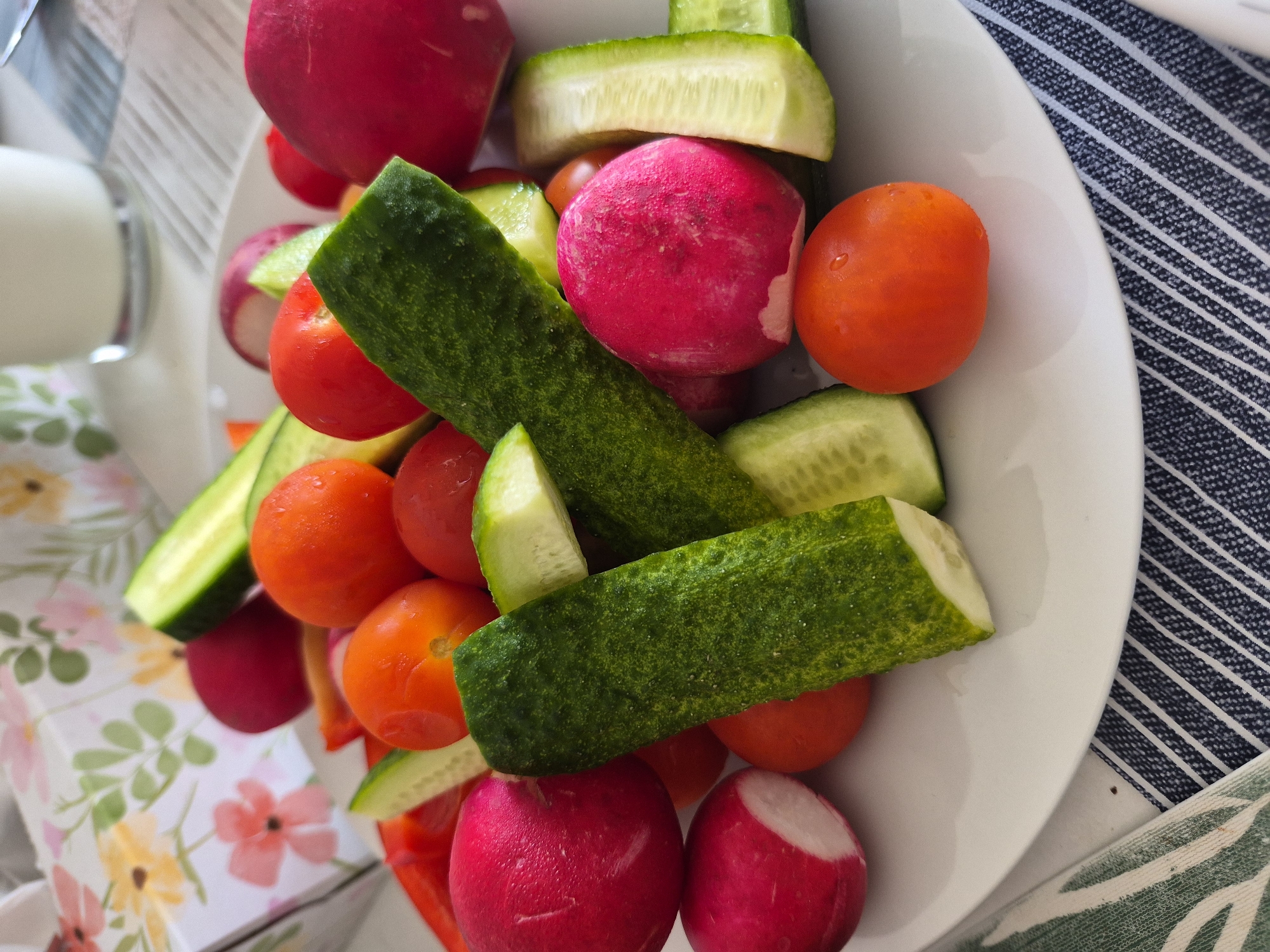A close-up, eye-level, full shot shows a white plate filled with sliced vegetables and some whole cherry tomatoes. The vegetables are arranged haphazardly, creating a layered effect. The plate is positioned on top of a striped blue and white napkin, with a green and white leaf-patterned cloth visible on the left side. In the background, a blurred floral-patterned box is visible, along with a glass of milk. The lighting is bright, suggesting it was taken indoors during the day.