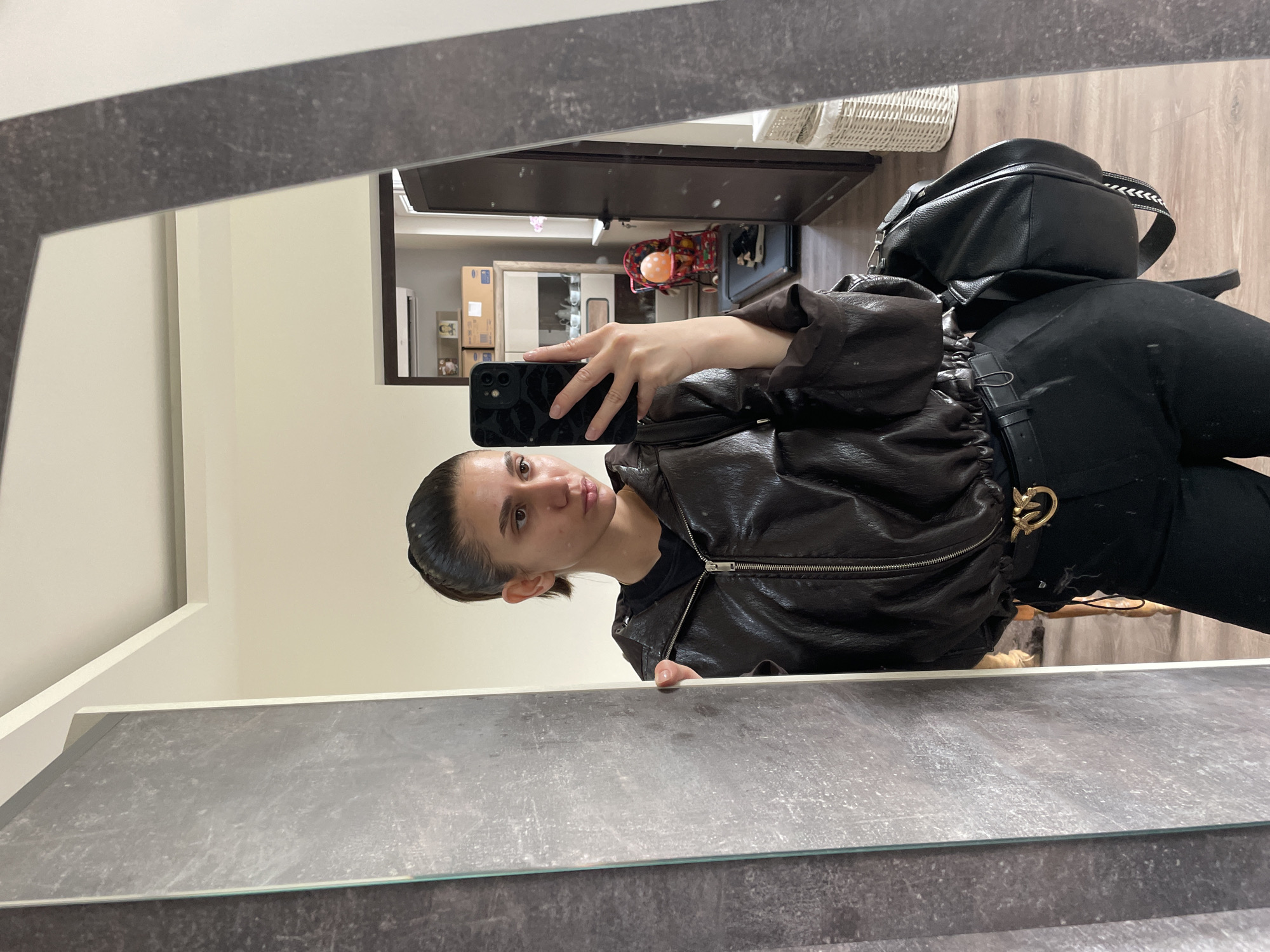 A young woman is taking a selfie in a mirror. She is wearing a brown leather jacket, black pants, and a black backpack. She has her hair pulled back and is making a kissing face. The background shows a hallway with a coat rack, furniture, and items stored on shelves. The setting appears to be an apartment interior, but there is no verifiable information to place this at Sofia, Bulgaria.