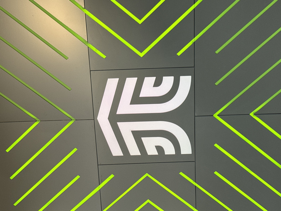 An indoor wall segment features a modern, geometric design. The surface consists of large, rectangular dark gray panels arranged in a grid. Centered on these panels is a distinct white, illuminated logo, composed of thick, rounded lines forming a stylized upward-pointing arrow or combined initialism, consistent with the Metrobús León (Sistema Integrado de Transporte - SIT) logo. Surrounding this central emblem, and extending across the entire visible wall, are numerous integrated neon green linear elements. These green lines are arranged diagonally, forming a repeating pattern of V-shapes and parallel strokes, acting as illuminated grooves within the panel structure. No individuals or actions are depicted. The scene is located within a station of the Metrobús León system in León, Guanajuato, Mexico.