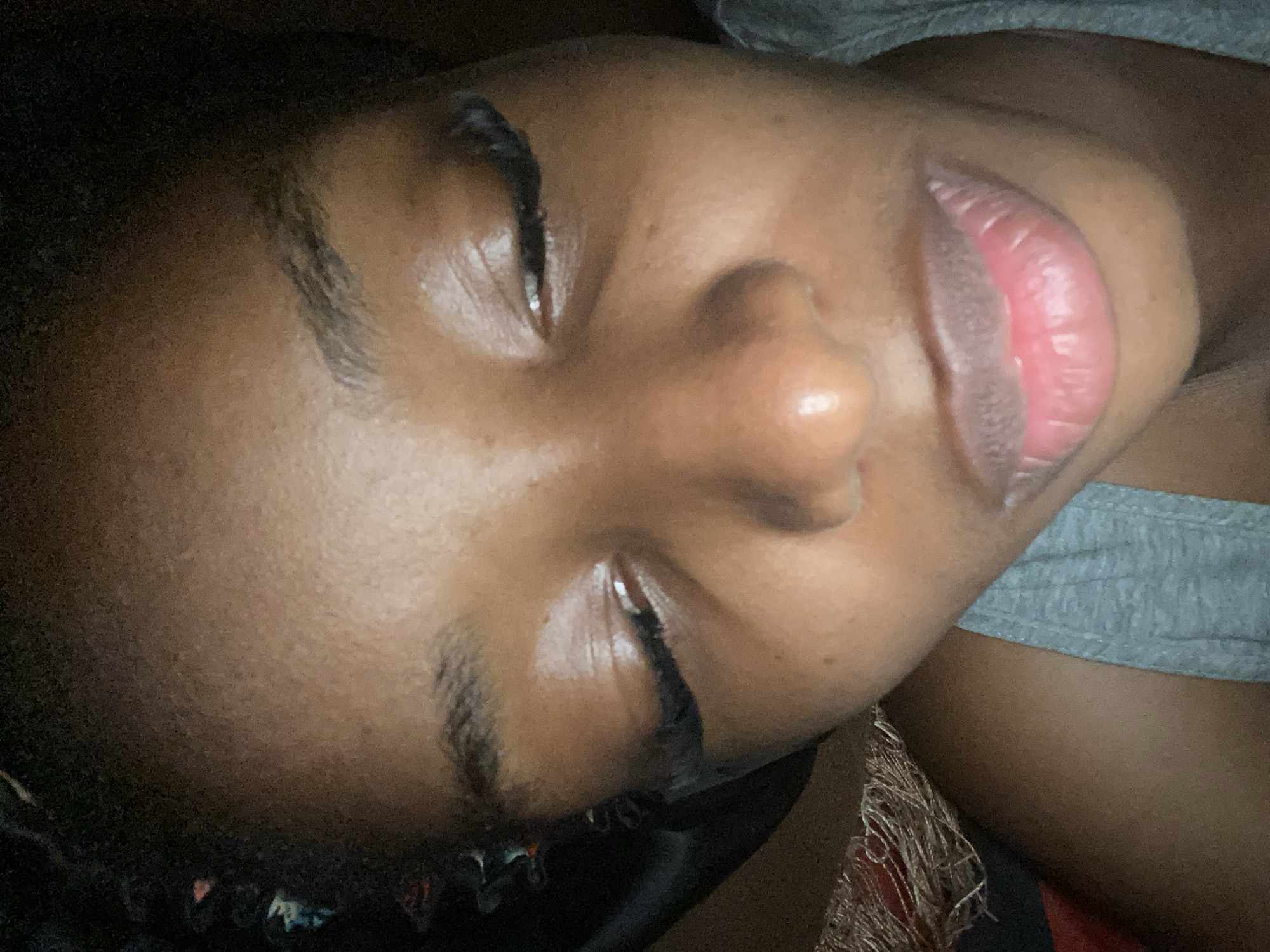 A close-up, low-angle, overhead shot shows the face of a dark-skinned person lying down. The person's eyes are partially closed, and they are wearing false eyelashes. The skin appears slightly oily, and there is makeup on the eyelids and lips. The person is wearing a grey tank top. The background is dark and appears to be indoors, possibly in a bedroom. There are no identifiable landmarks or contextual clues to indicate the location in Jos, Nigeria.
