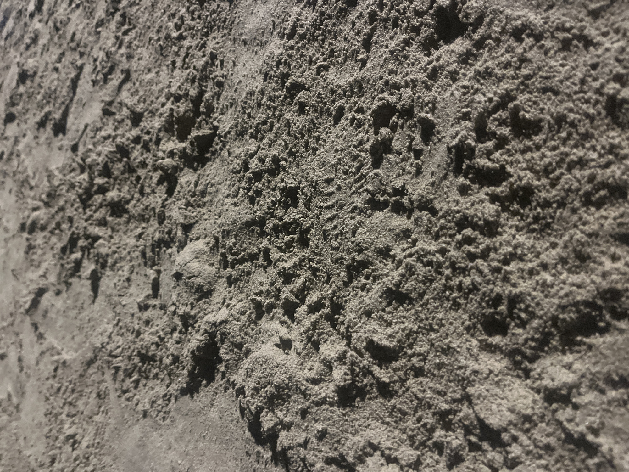 The image shows a close-up view of a surface covered in light-colored, granular material, resembling sand or fine gravel. The texture is rough and uneven, with small mounds and depressions casting shadows. The lighting suggests a strong light source from the left, creating a contrast between illuminated areas and shadowed regions. The surface appears to be part of the ground or a construction material. There are no people or discernible objects other than the granular material itself. The location is identified as Unknown, Unknown.