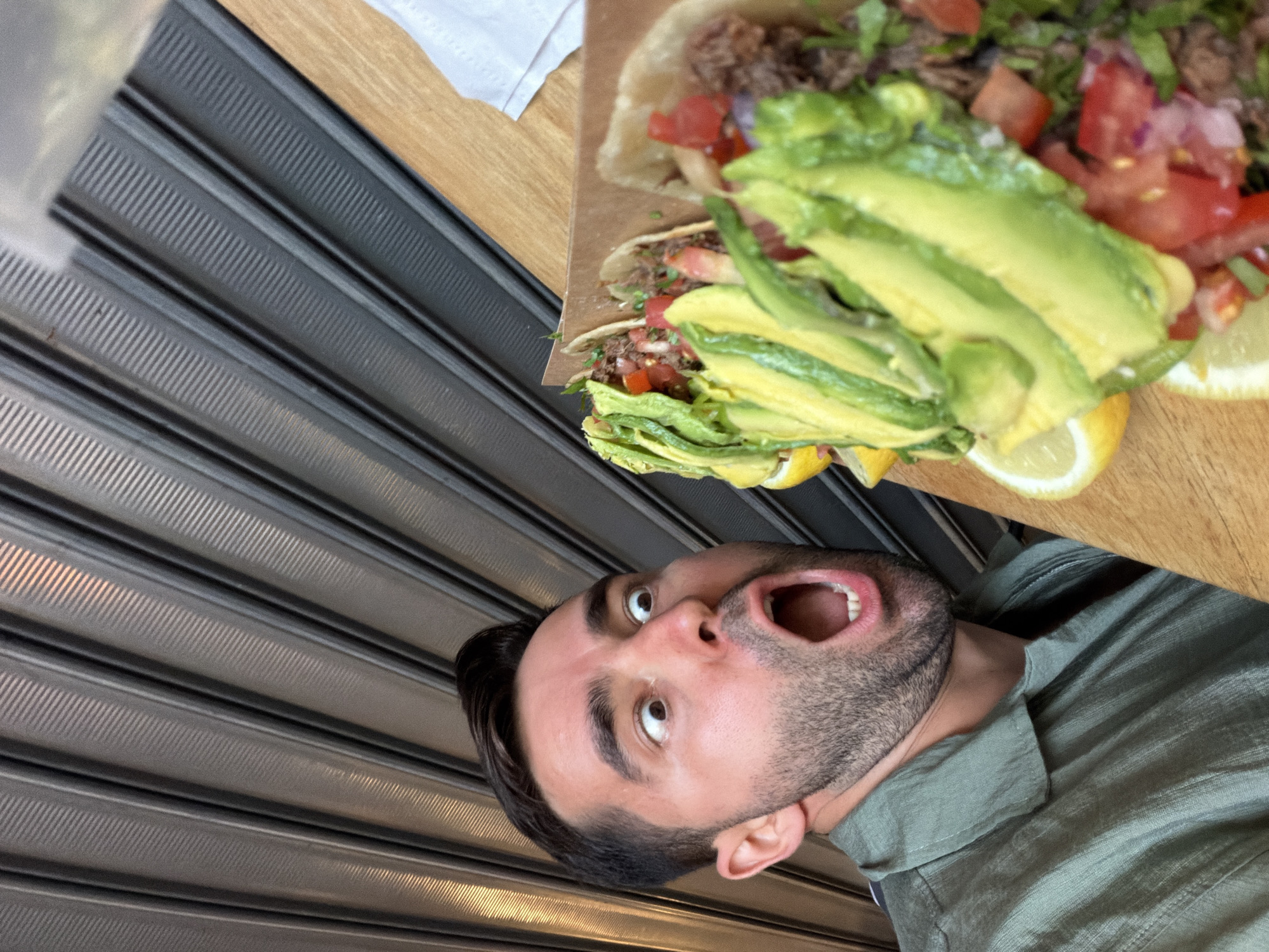 A man with dark hair and a beard is captured in an expressive moment, looking upwards with his mouth wide open in surprise or excitement. He is wearing a green collared shirt. The image is oriented with the man appearing vertically on the right side of the frame.
Adjacent to the man, on a light wooden surface, are two generously filled tacos. The tacos contain a filling of ground meat, topped with ample slices of fresh green avocado, diced red tomatoes, and finely chopped herbs. Several bright yellow lemon or lime wedges are placed around the tacos, suggesting they are meant for garnish or added flavor. A crumpled white napkin is also visible on the wooden surface in the upper left.
The background features a textured wall composed of distinct horizontal, ribbed metal panels, possibly a roll-down shutter or decorative metal cladding. The lighting is soft and even, indicating a daytime setting, likely indoors or under a covered outdoor area. The man's expression conveys strong anticipation or delight directed towards the food. The scene depicts a casual dining experience, possibly in Tel-Aviv, Israel. No visible text is present in the image.