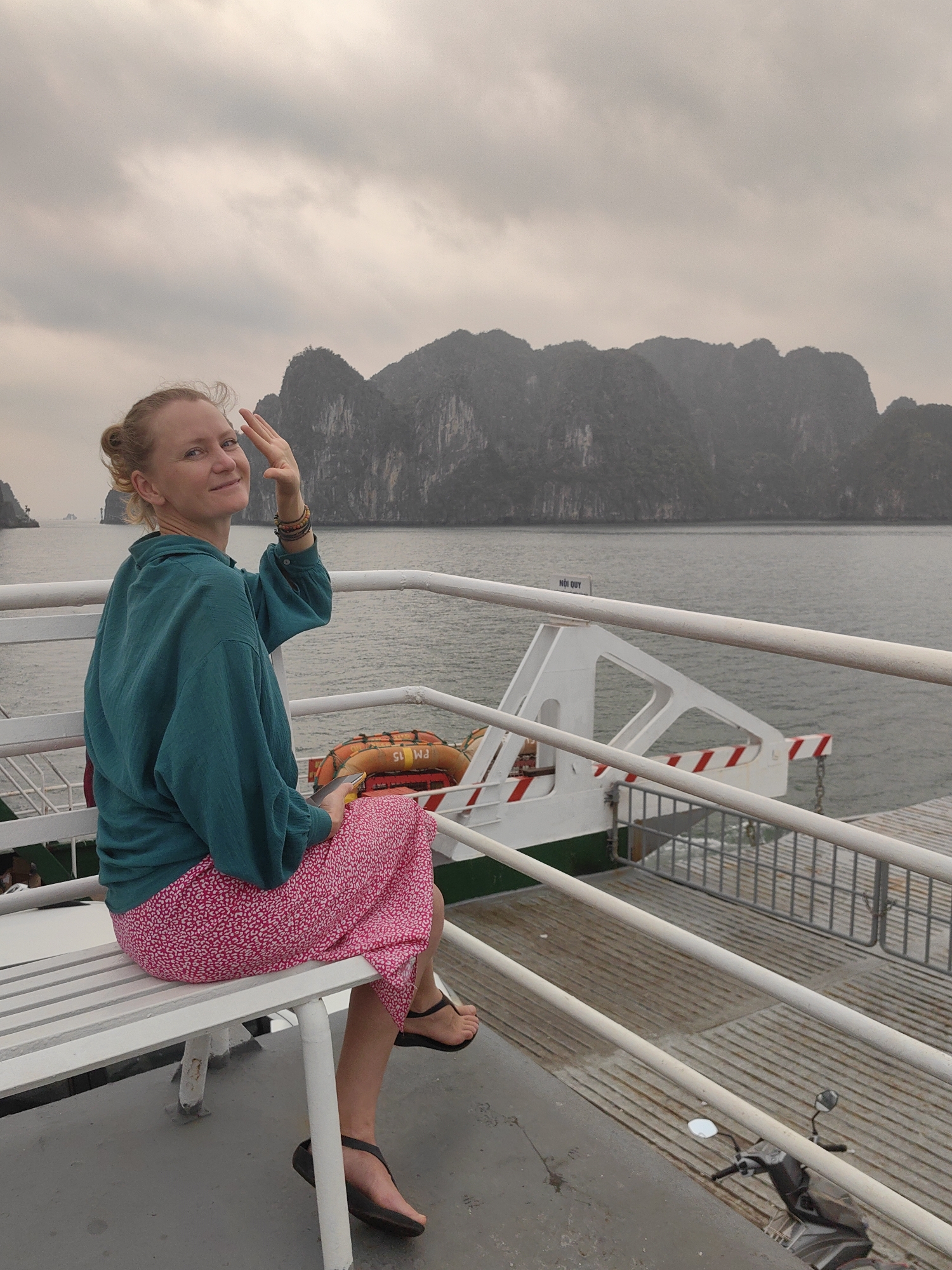 A medium shot depicts a blonde woman seated on a white bench on a ferry. The ferry appears to be in Tuan Chau Ward, Vietnam, given the landscape of limestone karsts in the background. The woman is wearing a teal, long-sleeved shirt and a knee-length, red and white floral skirt. She is turned towards the camera, smiling and waving with her right hand. She has bracelets on her right wrist. She is wearing black sandals. A small sign with Vietnamese writing is attached to the ferry. There are orange life preservers attached to the white ferry frame. A motorcycle is parked on the ferry deck. The sky is overcast, and the water is grey.