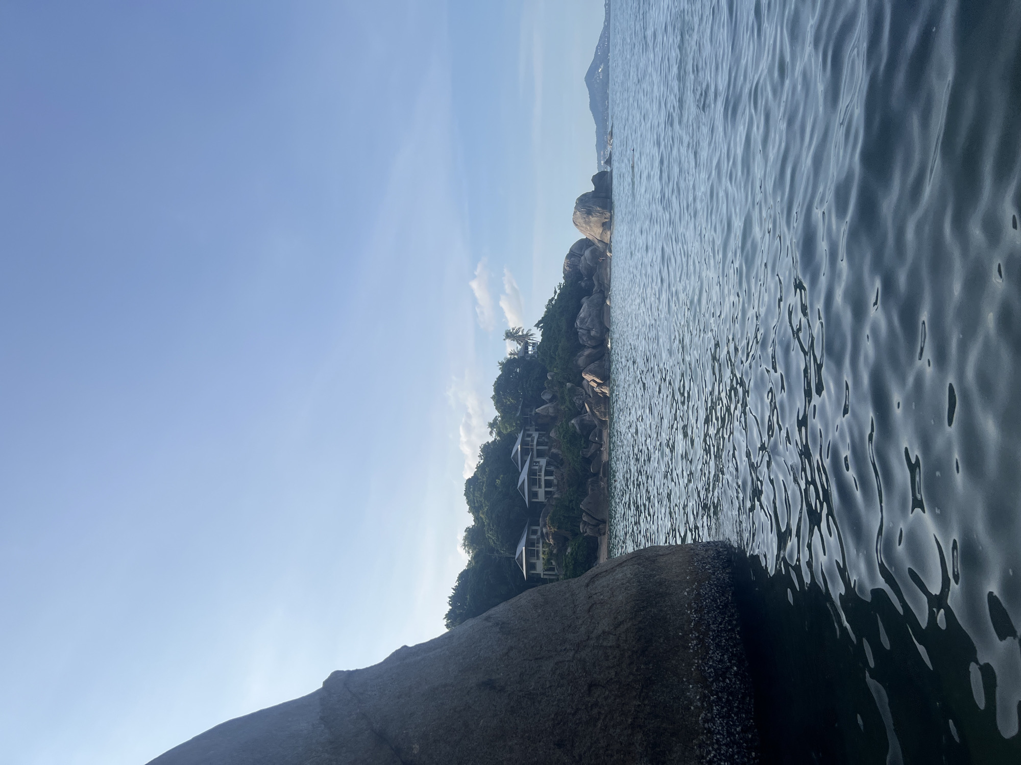 An eye-level shot captures a section of the water in Ko Samui District, Thailand, on a bright, clear day. The foreground is dominated by the dark, rippling surface of the sea, reflecting the sky and creating a dappled pattern of light and shadow. To the left, a massive, dark gray boulder juts out from the water, its base encrusted with barnacles or similar marine growth.
In the mid-ground, a lush, green hillside slopes down towards the water. Several white, modern villas with dark roofs are nestled among the trees, appearing to be part of a resort or private residences. The hillside is dotted with large, rounded boulders, and a small beach or clearing is visible between the rocks and the water. Palm trees and other tropical foliage cover the higher slopes.
The background features a vast expanse of a light blue sky with a few wispy white clouds. To the far right, a hazy mountain range or island is visible in the distance, suggesting the presence of other landmasses. The overall scene evokes a sense of tropical tranquility and luxury. No people are clearly identifiable in the image.
