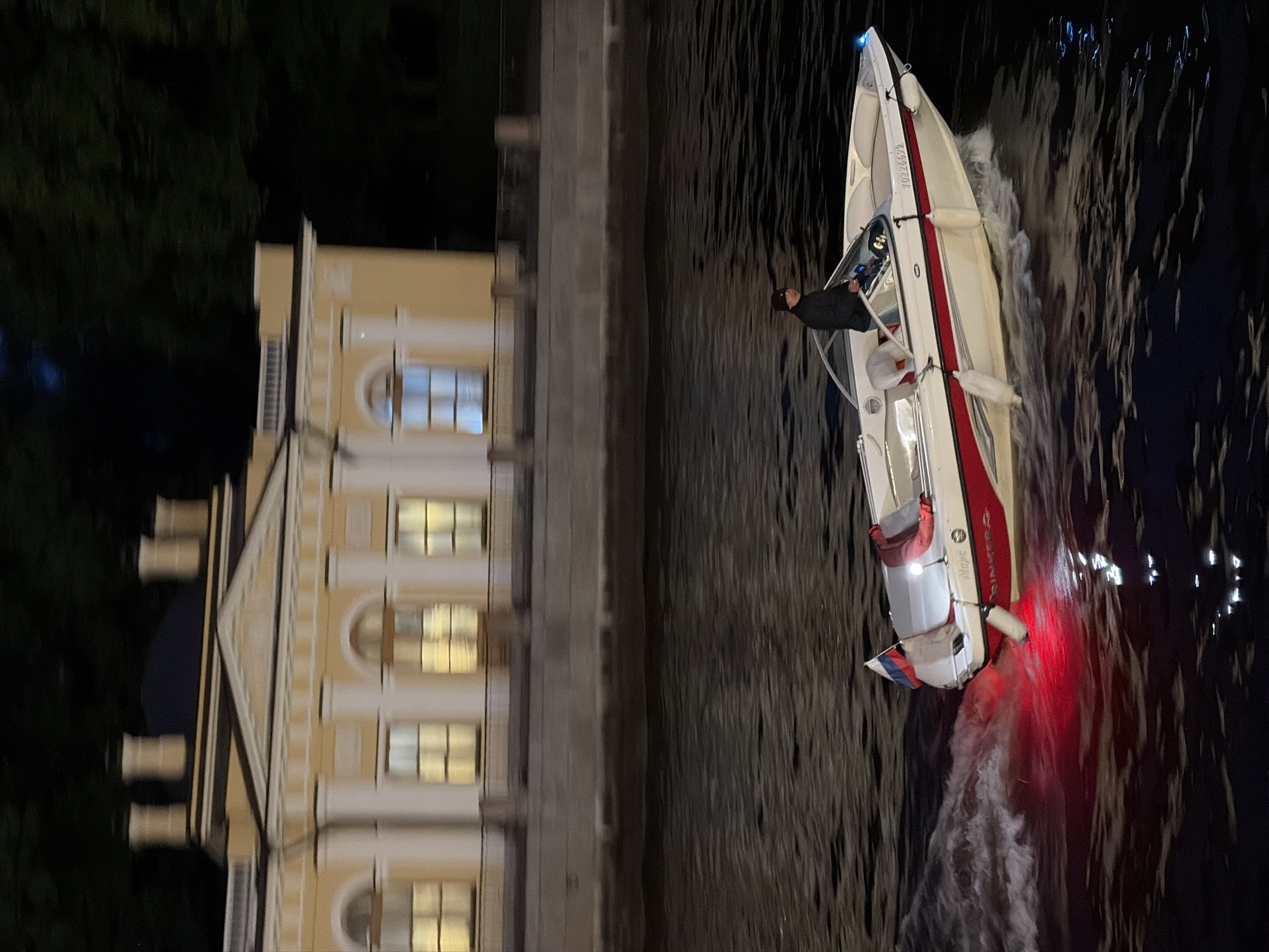 A motorized white and red boat with a Russian flag is moving at speed on a dark body of water, leaving a wake. A person in a black jacket and hat is at the helm of the boat. The boat is adorned with white fenders and has the registration number "1022609" visible on its side. Red lights are visible at the stern of the boat, illuminating the water. In the background, across the water, is a long, pale yellow building with a light-colored roof. The building features multiple windows, some of which are illuminated with warm light. The facade of the building has classical architectural elements, including columns and decorative detailing. Dark trees are visible behind the building, suggesting a nighttime scene in a city, likely St. Petersburg given the architectural style and the presence of a waterway and a building of this appearance.