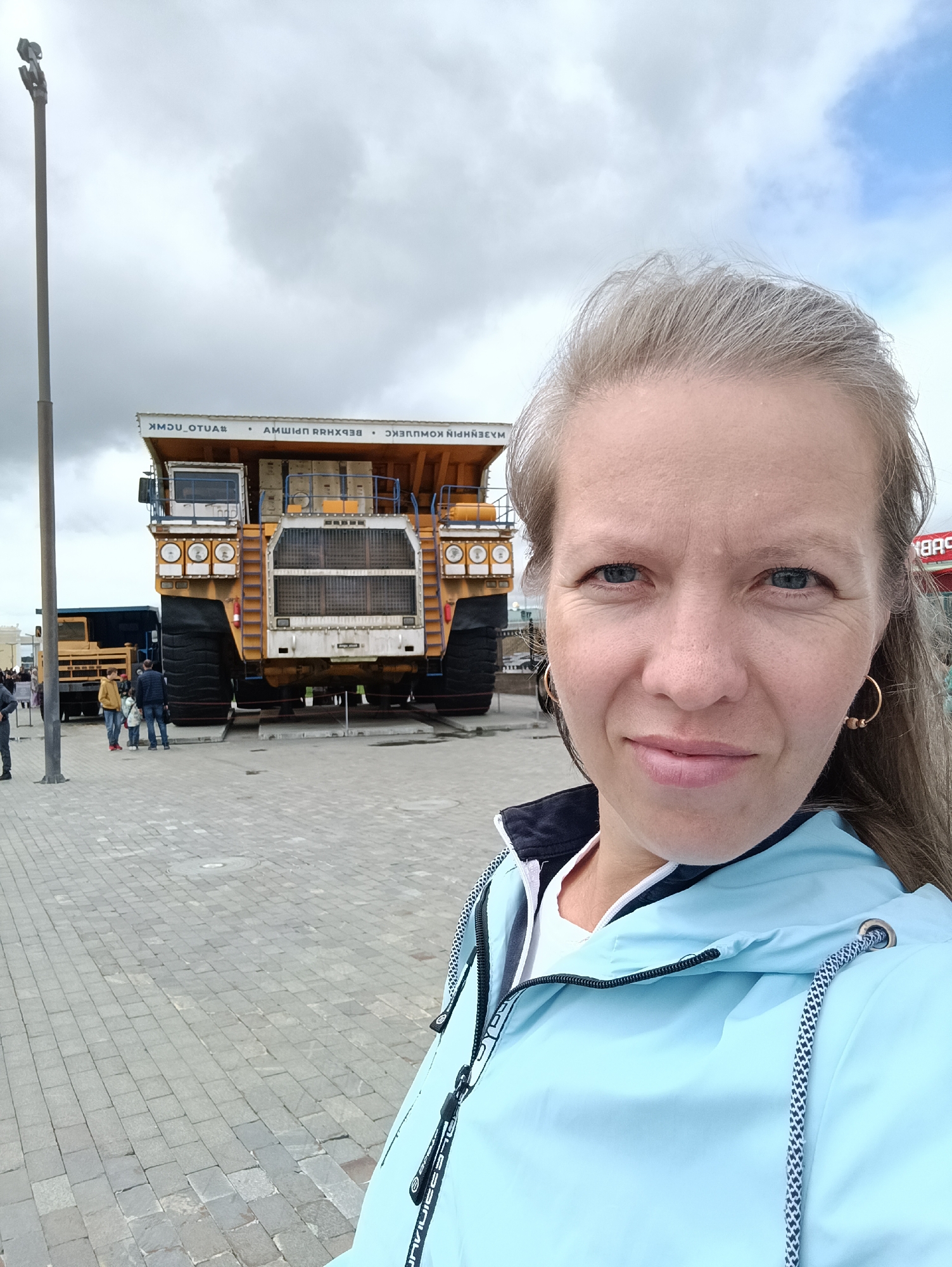 This is a medium shot selfie taken outdoors, featuring a woman in the foreground and a large mining truck in the background. The woman, who appears to be in her late 30s or early 40s, has light skin, blue eyes, and shoulder-length grey-blonde hair. She is wearing a light blue hooded jacket with a dark collar over a white top. She has hoop earrings in both ears. The woman is smiling slightly and looking at the camera.
Behind her, a colossal yellow mining truck is parked on display. The truck is enormous, with massive tires and a large cabin. Its front grille and headlights are visible, as well as part of its cargo bed. The truck is elevated on platforms, suggesting it is a museum piece or monument. To the left of the main truck, another, smaller dark blue truck is partially visible.
The setting is an outdoor plaza paved with grey interlocking stones. A tall lamppost stands to the left of the frame. In the distance, a few other people can be seen, appearing as small figures relative to the trucks. The sky is cloudy with patches of blue.
The visible text on the truck, oriented horizontally and likely in Cyrillic script, reads: "ВЕРХНЕПЫШМИНСКИЙ КОМПЛЕКС" which translates to "VERKHNEPYSHMINSKY COMPLEX" and "ОТЦА" which is likely part of a longer word or phrase. There is also some text on the woman's jacket, appearing to be a brand name in a stylized font.
The overall impression is of a visit to an industrial or mining museum in Verkhnyaya Pyshma, Russia, on a day with mixed weather. The woman appears happy and engaged with her surroundings.
