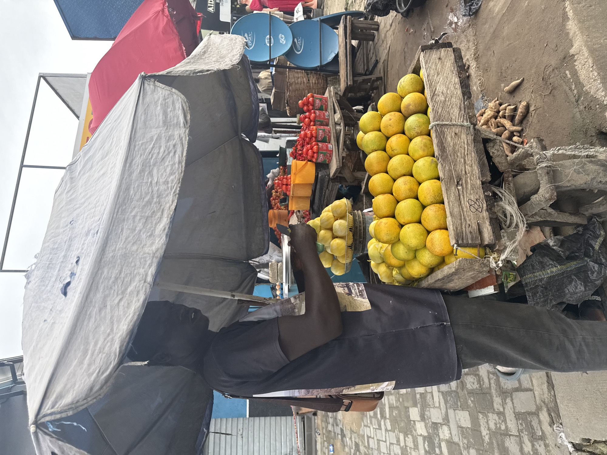 A man stands under a tattered grey umbrella at a street market in Ado, Nigeria. He is wearing a dark t-shirt with a graphic print on the back and dark pants. He is looking to his right and holding a mobile phone in his right hand. In front of him, a wooden crate is overflowing with ripe, yellow-green oranges. To the left of the oranges, a smaller pile of lighter yellow fruits, possibly lemons, is visible. Behind the oranges, a wooden table is laden with red tomatoes in cans and yellow buckets. Further back, blue satellite dishes are mounted on a structure, and a woman in a red dress is visible. The ground is paved with grey and white bricks, and other market stalls and produce are visible in the background. The overall scene depicts a vibrant street market environment.