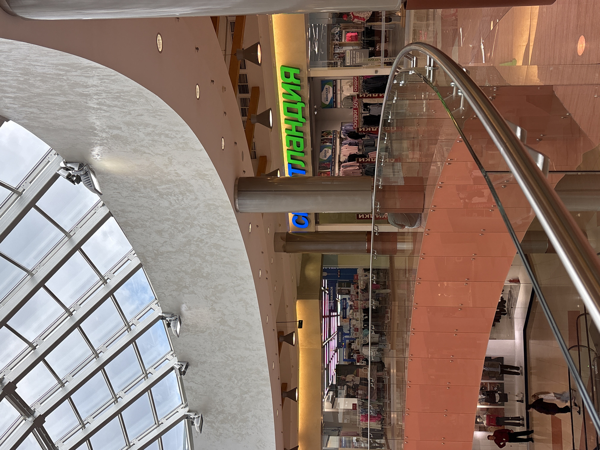 This image shows the interior of a shopping mall. A large, curved glass and metal railing system dominates the foreground, following a ramp or staircase. In the background, there are shops with brightly lit displays and clothing racks. One shop sign prominently features green lettering that spells "ТЛАНДИЯ", which is Russian for "Land of..." or "Tlandia". Another shop has a sign with blue lettering. The ceiling is a large, multi-paneled glass skylight, allowing natural light to filter into the space. There are also artificial lights visible, including modern spotlights attached to the ceiling structure and pendant lights hanging over the shop entrances. People are visible on different levels of the mall, some walking and others standing near shop displays. The architecture appears modern, with smooth, textured walls and large support columns. The overall impression is of a busy, well-lit commercial interior. It is not possible to determine the city of Perm, Russia, from the image itself, but based on the Russian text on the signage, it is likely a Russian-speaking country.