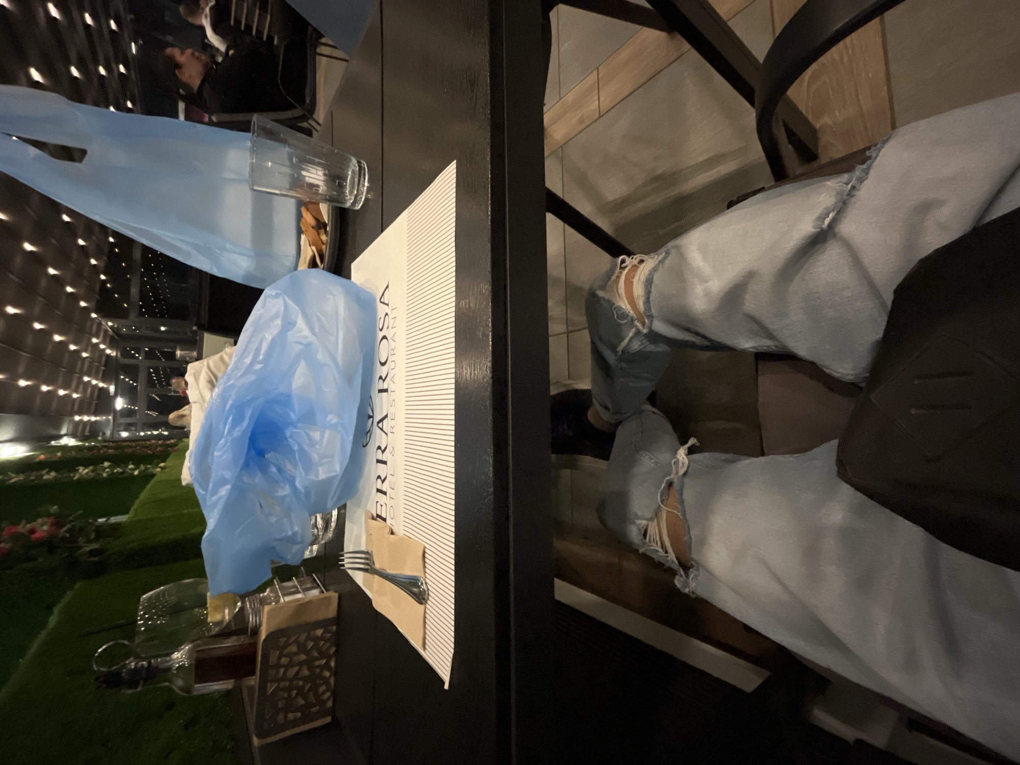 The image depicts a dining scene, likely in a restaurant. A person is seated at a table, wearing ripped light-wash jeans and a black bag on their lap. The table setting includes a branded paper placemat for Terra Rosa Hotel & Restaurant, glassware, a fork, a napkin, and a bottle of red wine. Blue plastic bags obscure the view of other patrons and the restaurant's interior, but the background shows a grassy area and what seems to be exterior lights, indicating an outdoor or partially open dining area. Considering the branding, the location is likely at or near the Terra Rosa Hotel & Restaurant in Karlovo, Bulgaria.