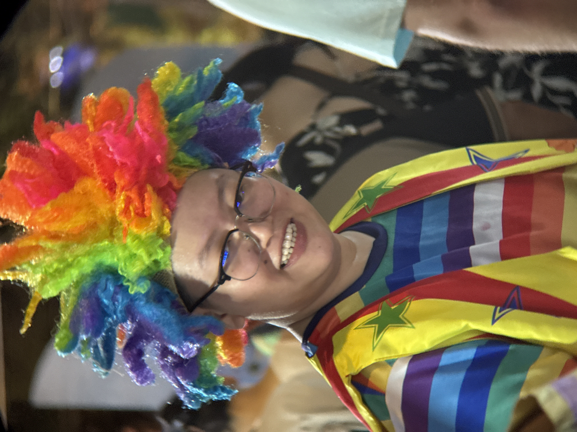 A person wearing a multi-colored wig and a rainbow-striped shirt and vest smiles at the camera. The person has short dark hair and wears glasses. The background is blurred, but appears to be an outdoor event with other people visible. The location is identified as Nha Trang, Vietnam.