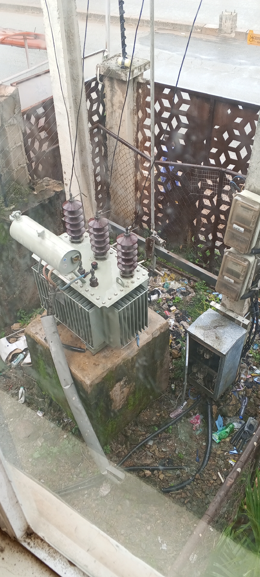 An outdoor scene depicts a pad-mounted transformer, a common electrical component, situated on a concrete base. The transformer is a light gray color with visible cooling fins and three dark brown ceramic insulators, each with multiple stacked discs. A light-colored cylindrical tank is mounted on top of the transformer. Adjacent to the transformer, a rusted metal enclosure, possibly for electrical equipment, is visible. The surrounding ground is littered with debris, including plastic bags, paper, and what appears to be vegetation. Several electrical wires are connected to the transformer and run across the scene, some supported by a concrete pillar and a fence made of diamond-shaped metal patterns. Two transparent electrical meters are mounted on a pole to the right of the transformer. The overall environment appears somewhat neglected and untidy, with evidence of litter and some overgrowth. The scene is captured from an elevated perspective, possibly from a window, indicated by a clear pane of glass in the foreground. The location is identified as Jos, Nigeria.