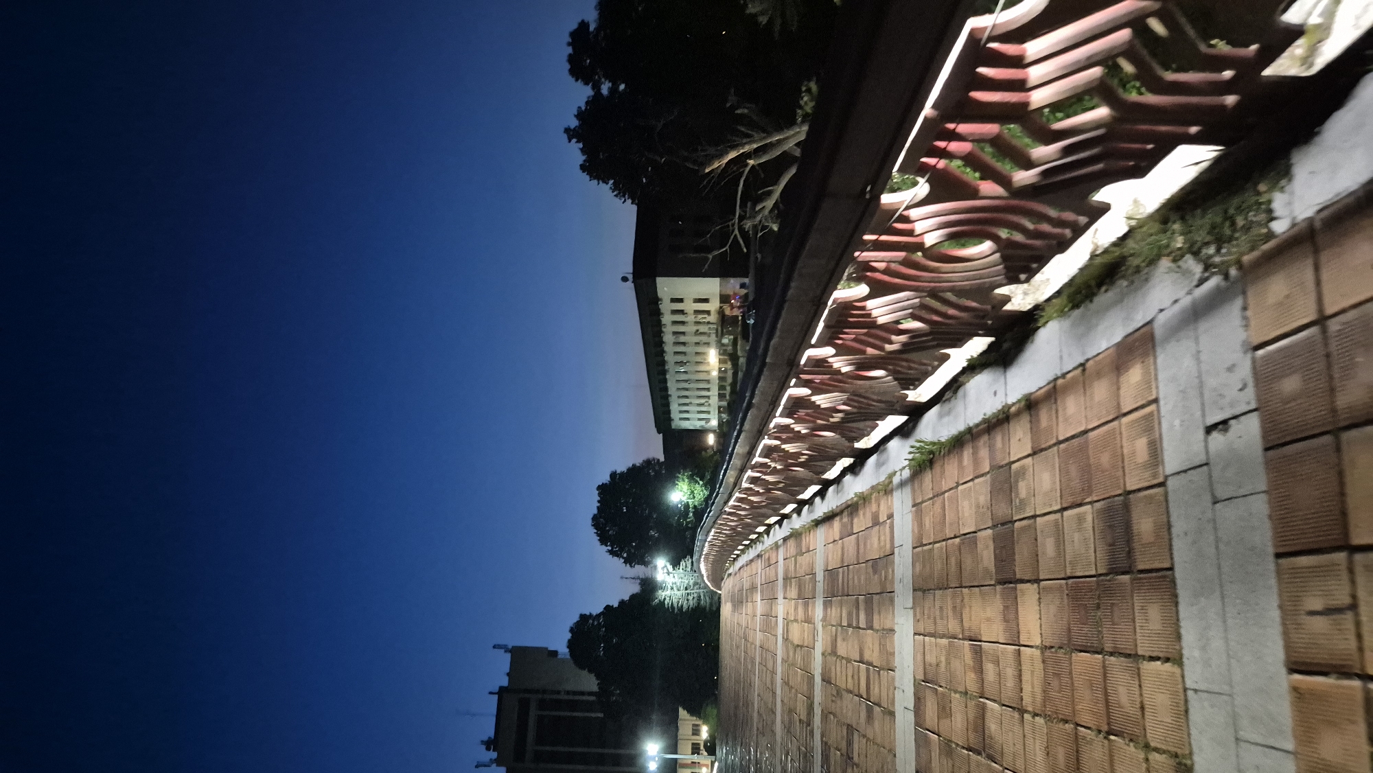 The image depicts a paved walkway with a patterned red railing, likely a bridge or elevated path, at dusk in Lovech, Bulgaria. The walkway is constructed of rectangular terracotta-colored tiles interspersed with light gray concrete borders. The railing features an intricate, decorative design with cutouts that are illuminated from within, casting a warm glow.
In the background, two prominent buildings are visible. The one on the left is a multi-story structure with visible antenna elements on its roof. The building on the right is a long, lighter-colored edifice with numerous windows. Trees, some appearing bare and others with foliage, are scattered throughout the scene, silhouetted against the darkening sky. Artificial lighting from street lamps illuminates the walkway and parts of the surrounding area. The sky is a deep twilight blue.
There are no identifiable people actively engaged in any discernible actions within the frame. The scene appears to be a quiet, public space captured at the transition from day to night. The architectural style of the buildings and the overall ambiance suggest an urban or institutional setting.