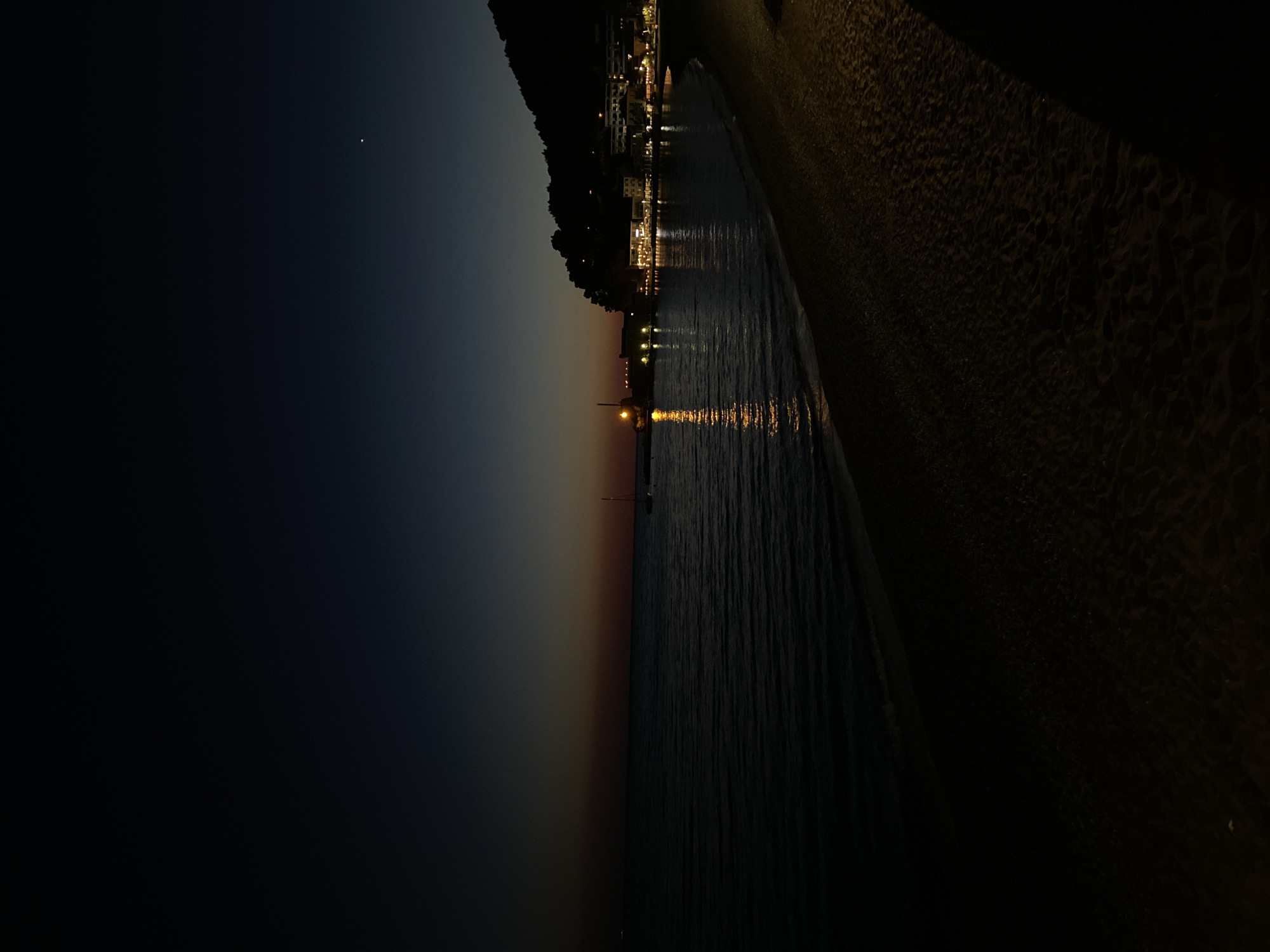 The image captures a serene coastal scene at twilight, transitioning into night, in Petrovac centar, Montenegro. The scene is oriented correctly to show the horizon line.
The dark sky, with a single bright star or planet visible in the upper left, indicates late evening. Along the horizon, a faint band of orange and red twilight colors suggests the recent sunset.
On the left side of the frame, a dark, tree-covered hill or mountain slopes down to the water's edge. At its base, a town is illuminated by numerous lights from buildings, which cast shimmering reflections across the calm sea. In the mid-ground, a pier or jetty extends into the water, marked by a prominent light at its end, creating a long, golden reflection on the water's surface. A faint outline of a sailboat mast can be seen near the pier.
The right side of the image features a dark, textured beach, likely sand or pebbles, illuminated softly by ambient light, showing subtle patterns in its surface. The water, slightly rippled, reflects the town and pier lights, creating elongated streaks of light.
The overall mood is peaceful and tranquil, characteristic of a quiet evening by the sea. No visible text is present in the media file.